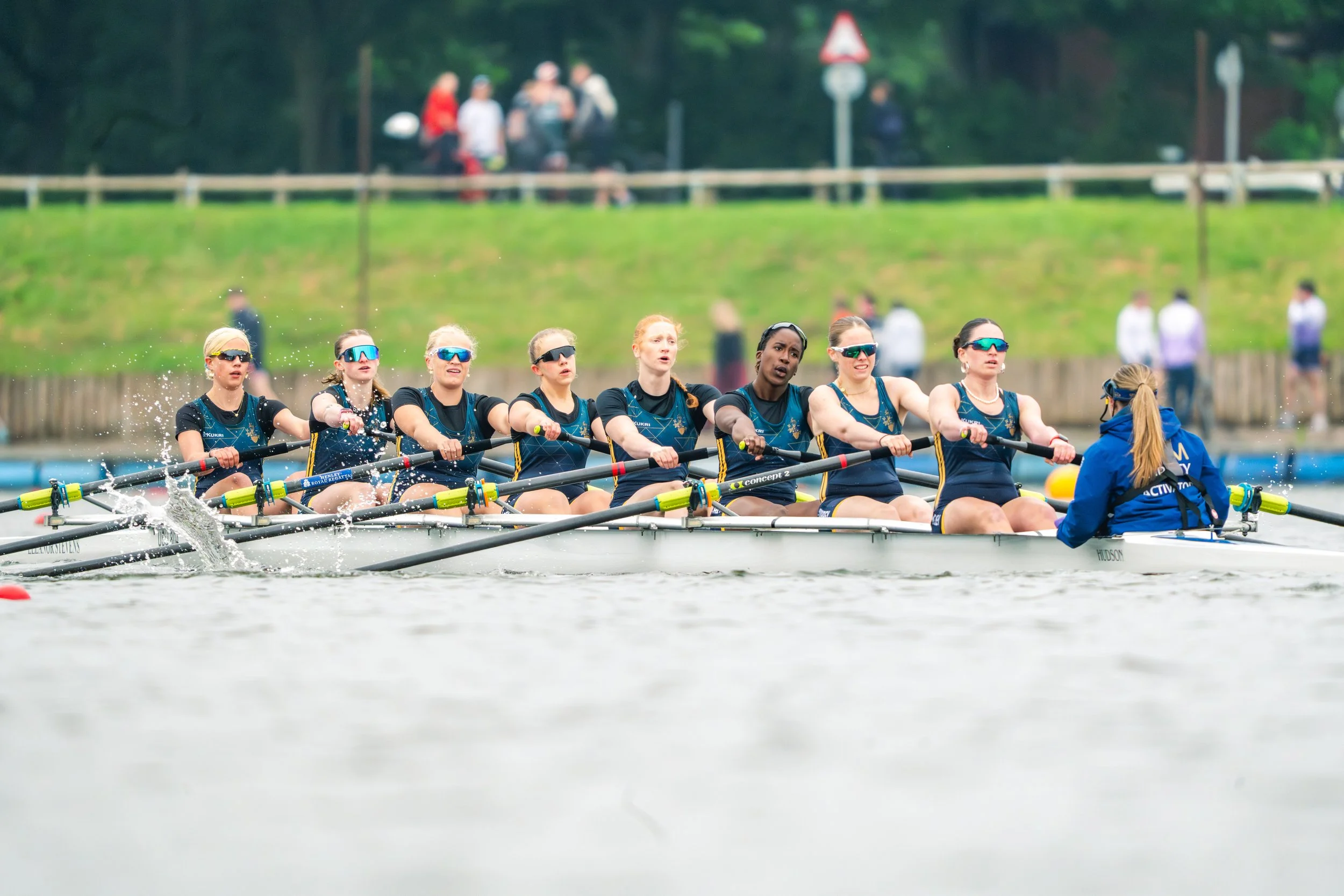 A group of women participating in a rowing race, sitting in a boat and rowing together on a body of water, with a green grassy area and a few spectators in the background.