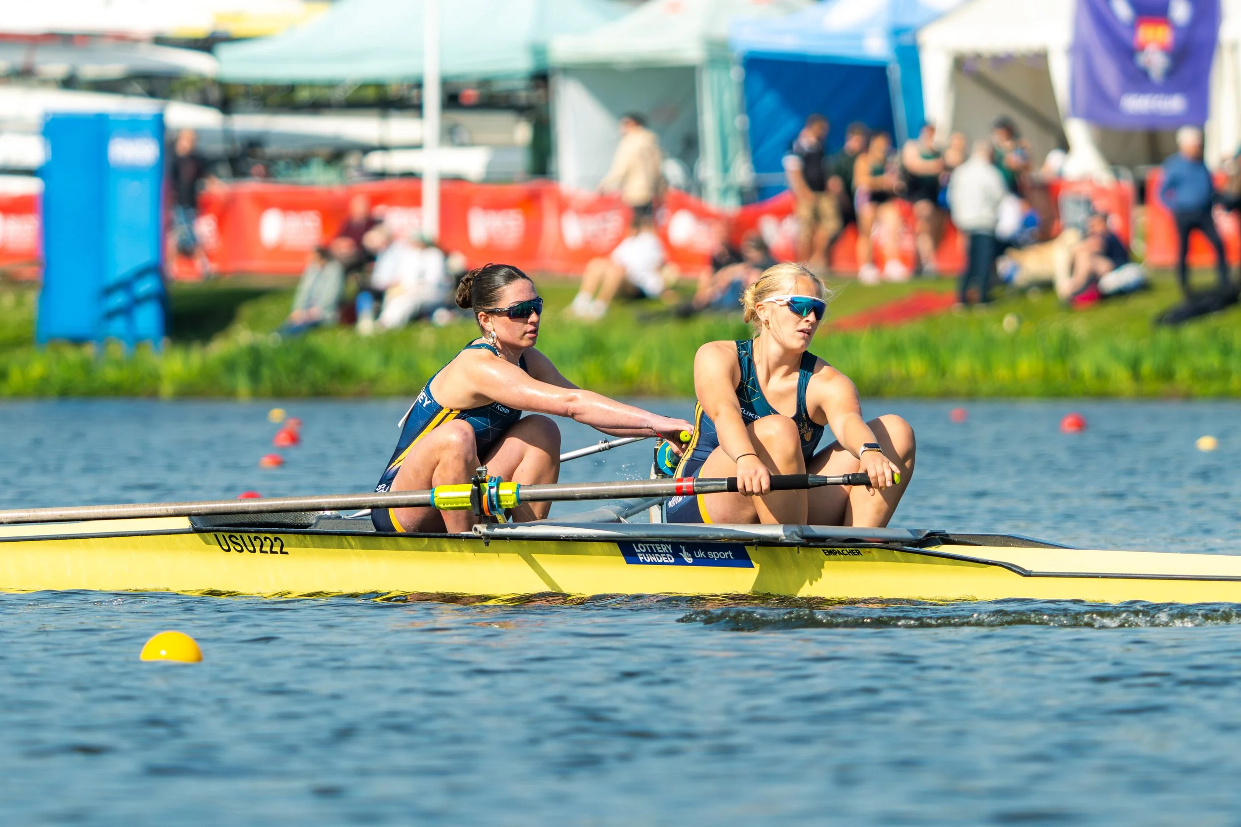 Two female athletes rowing a yellow boat during a race in a body of water, with spectators and tents in the background.