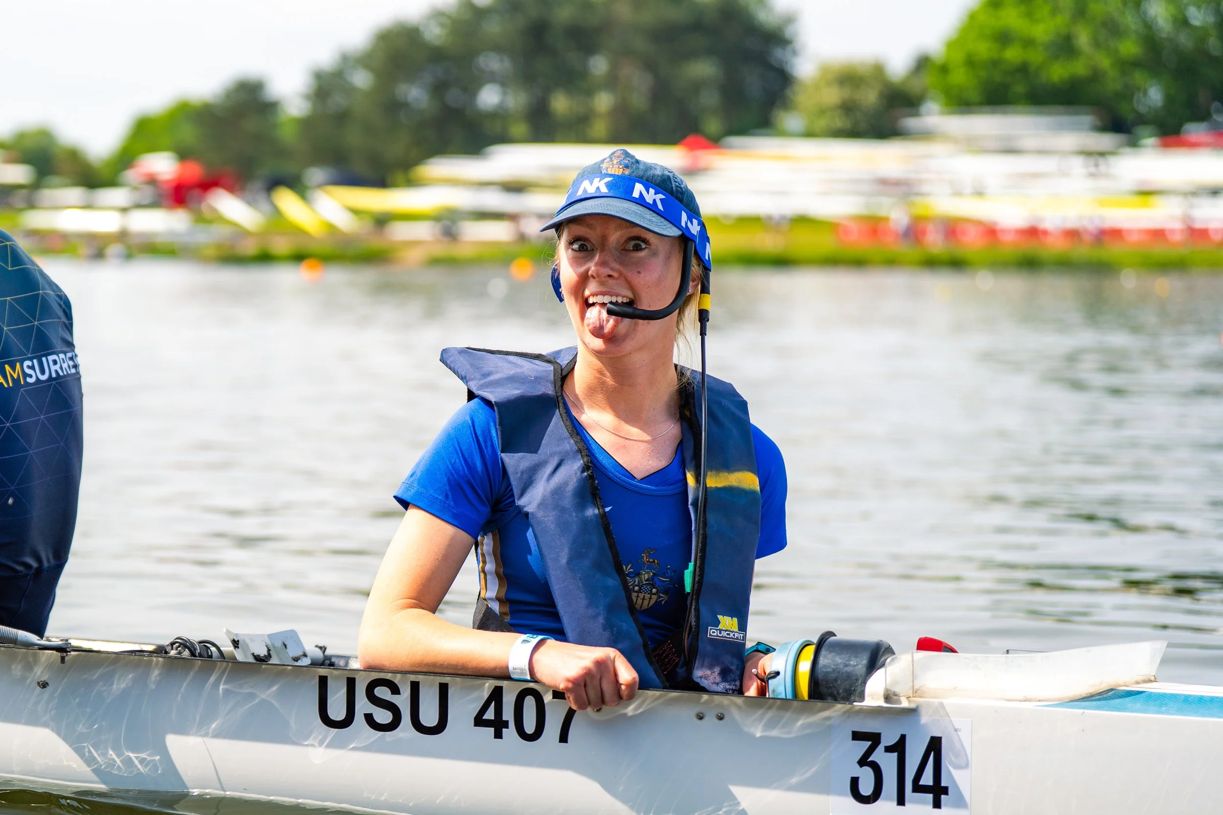 Woman in blue shirt and life jacket making a funny face with her tongue out while sitting in a small boat on a body of water, with blurred trees and boats in the background.