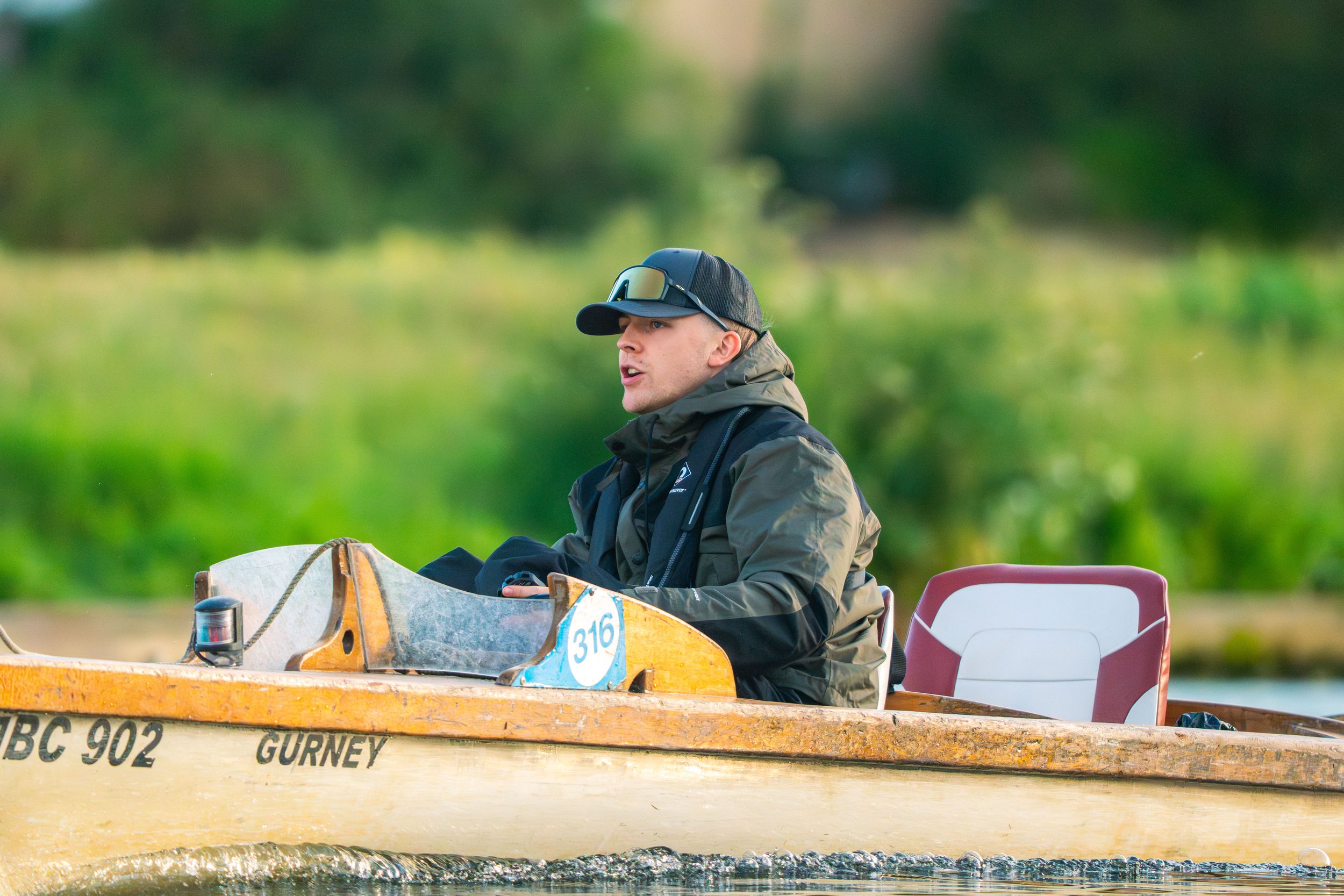 A man wearing a black cap, sunglasses on top, and a dark jacket sitting in a yellow boat labeled '316 Gurney' on a body of water. The background is blurred greenery.
