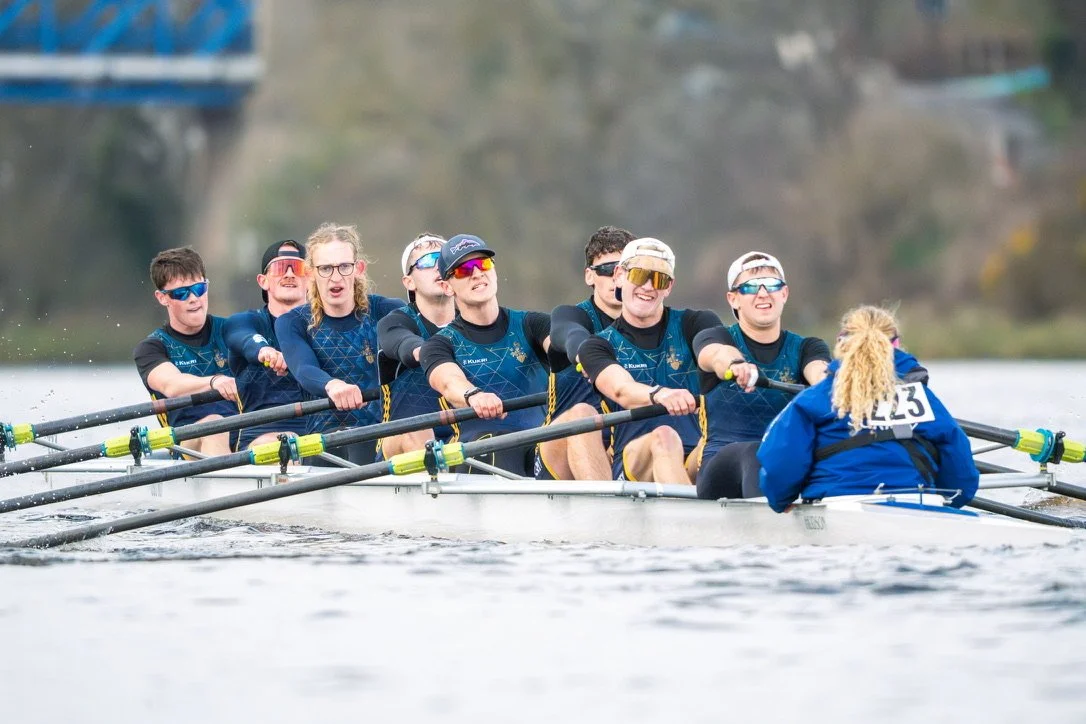 A team of eight rowers in blue and black uniforms rowing a boat on a river, with a person in a blue jacket at the front steering.