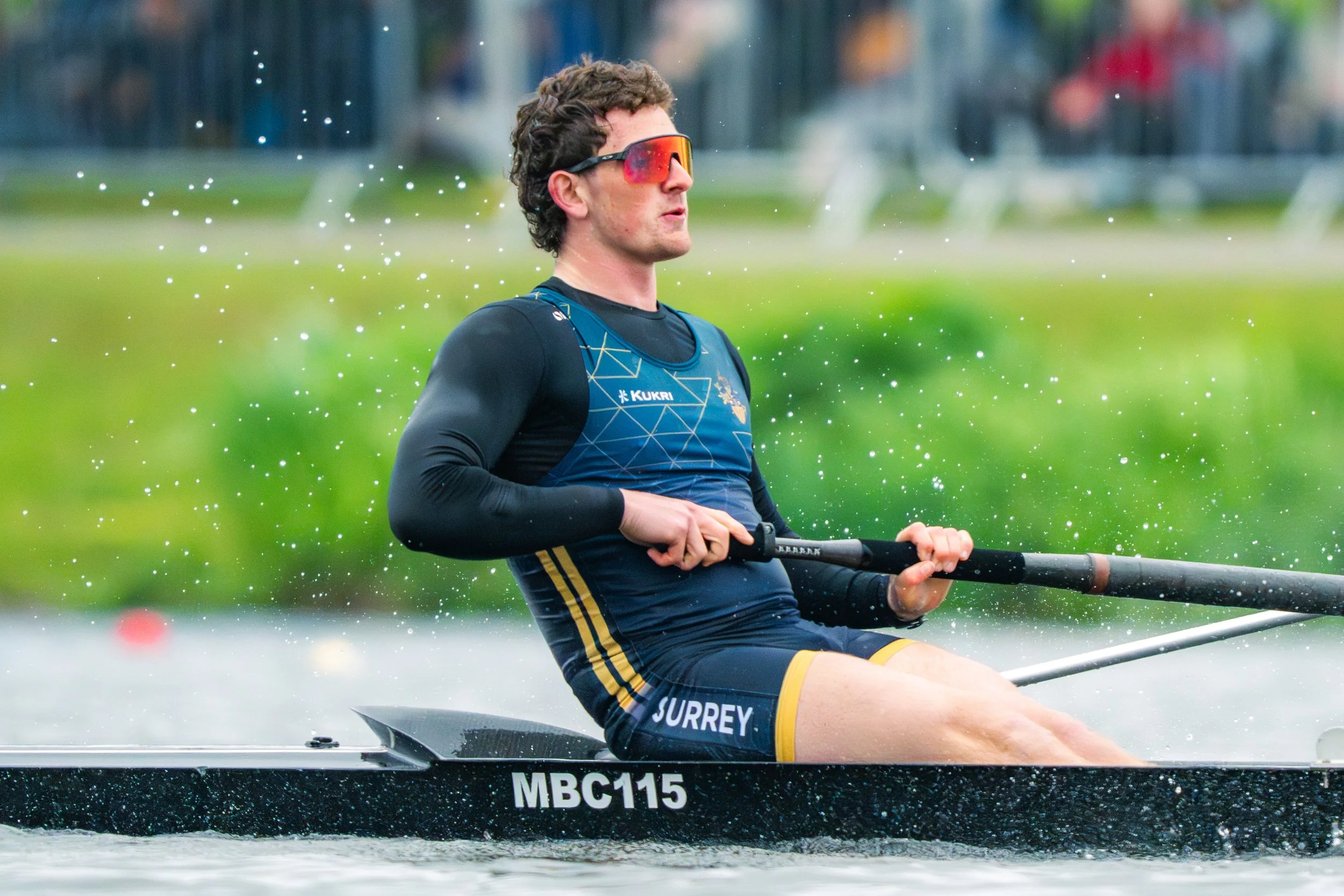 A male athlete in sports gear and sunglasses rowing a boat on water, with green trees and a blurred background.