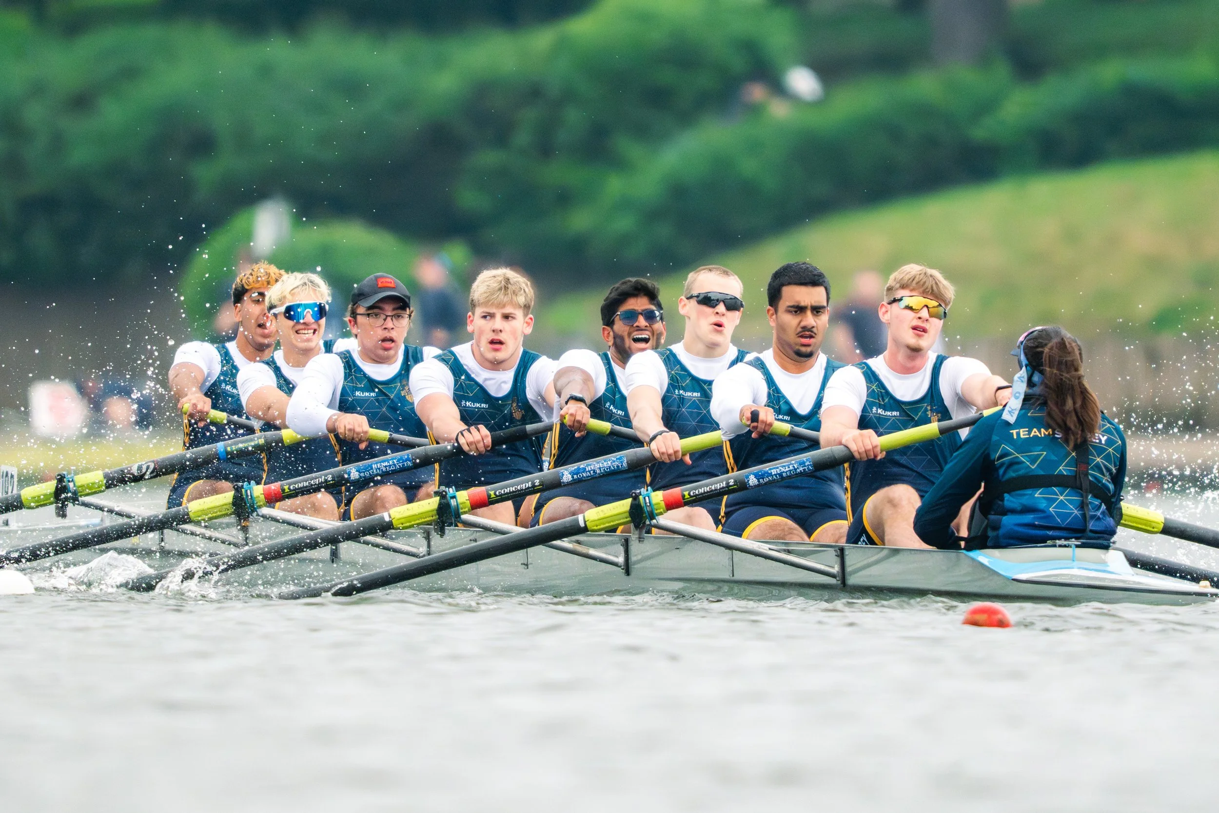 A team of young rowers competing in a boat race on a river, wearing sports uniforms and sunglasses, with a person at the front steering.