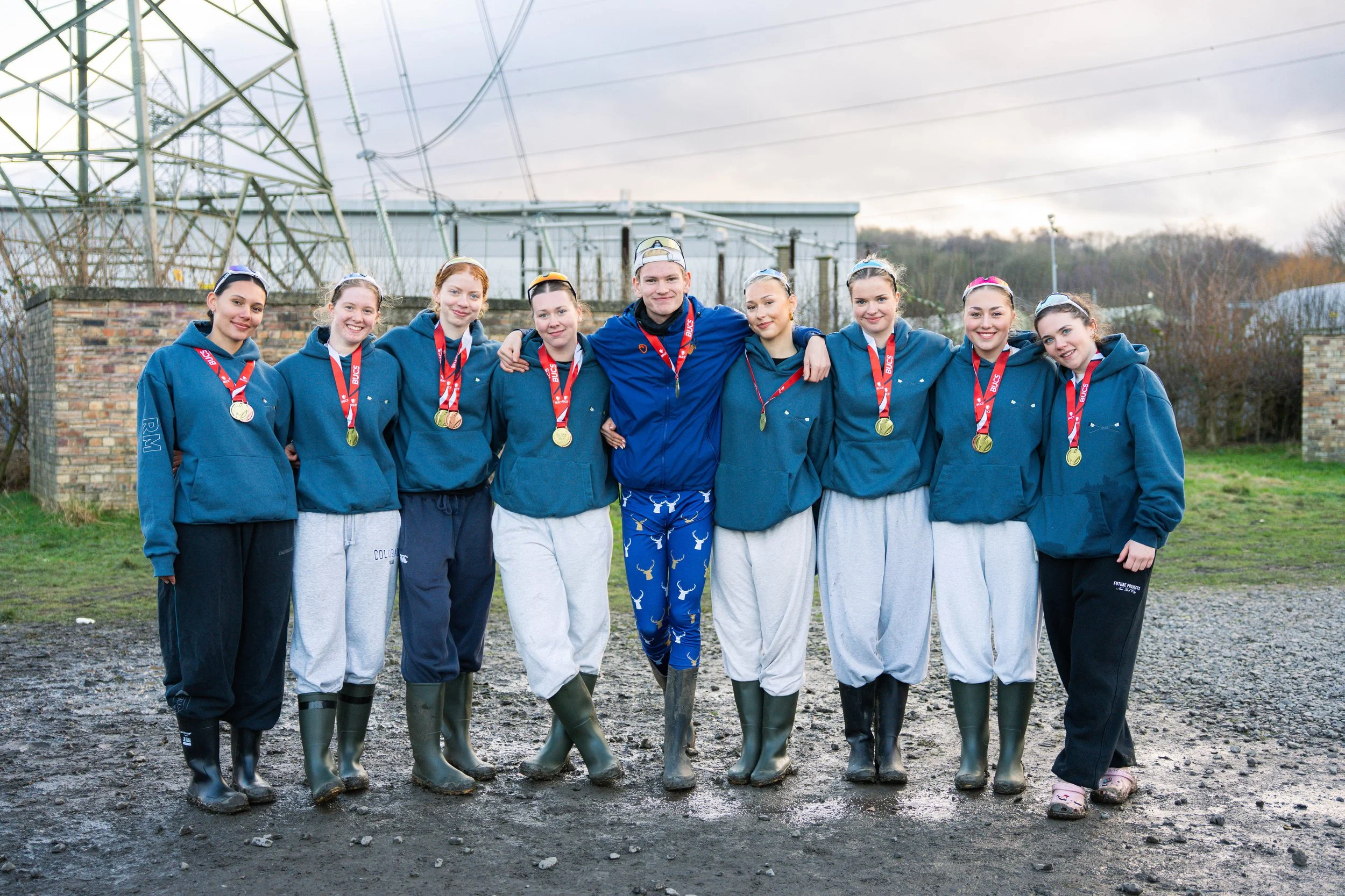 A group of eight young women and one young man standing outdoors in front of power lines and a brick wall, all wearing casual clothing including hoodies, sweatpants, and rain boots. They are smiling and wearing medals around their necks.