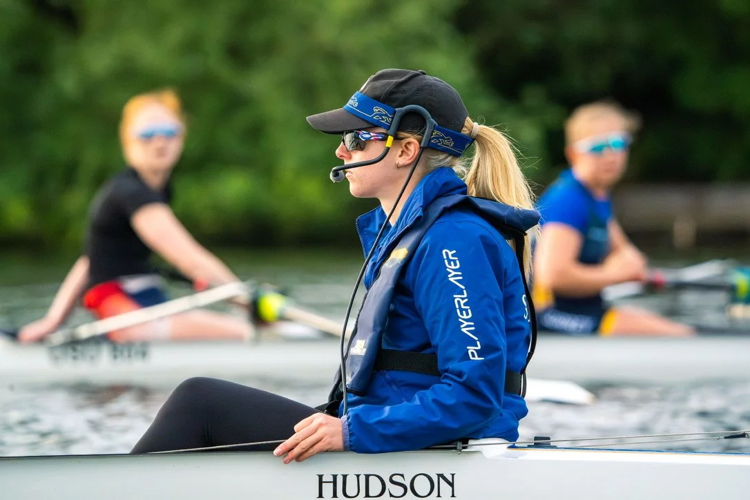 A woman wearing sunglasses, a black cap, and a blue jacket with a headset sitting in a boat named HUDSON, with two other women rowing in the background on a lake.