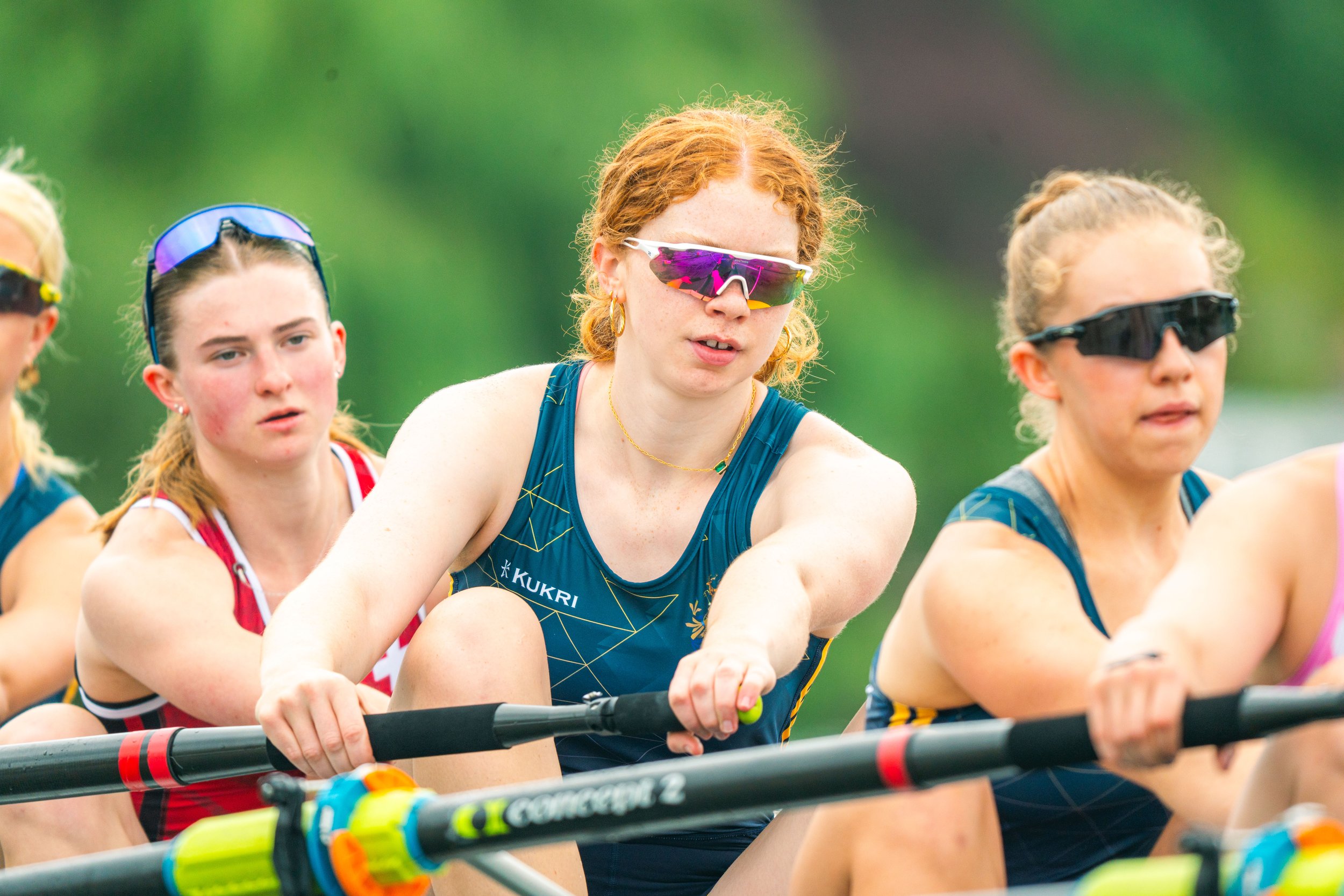 Rowing team in action, wearing athletic gear and sunglasses, holding oars during a race. Green blurred background.