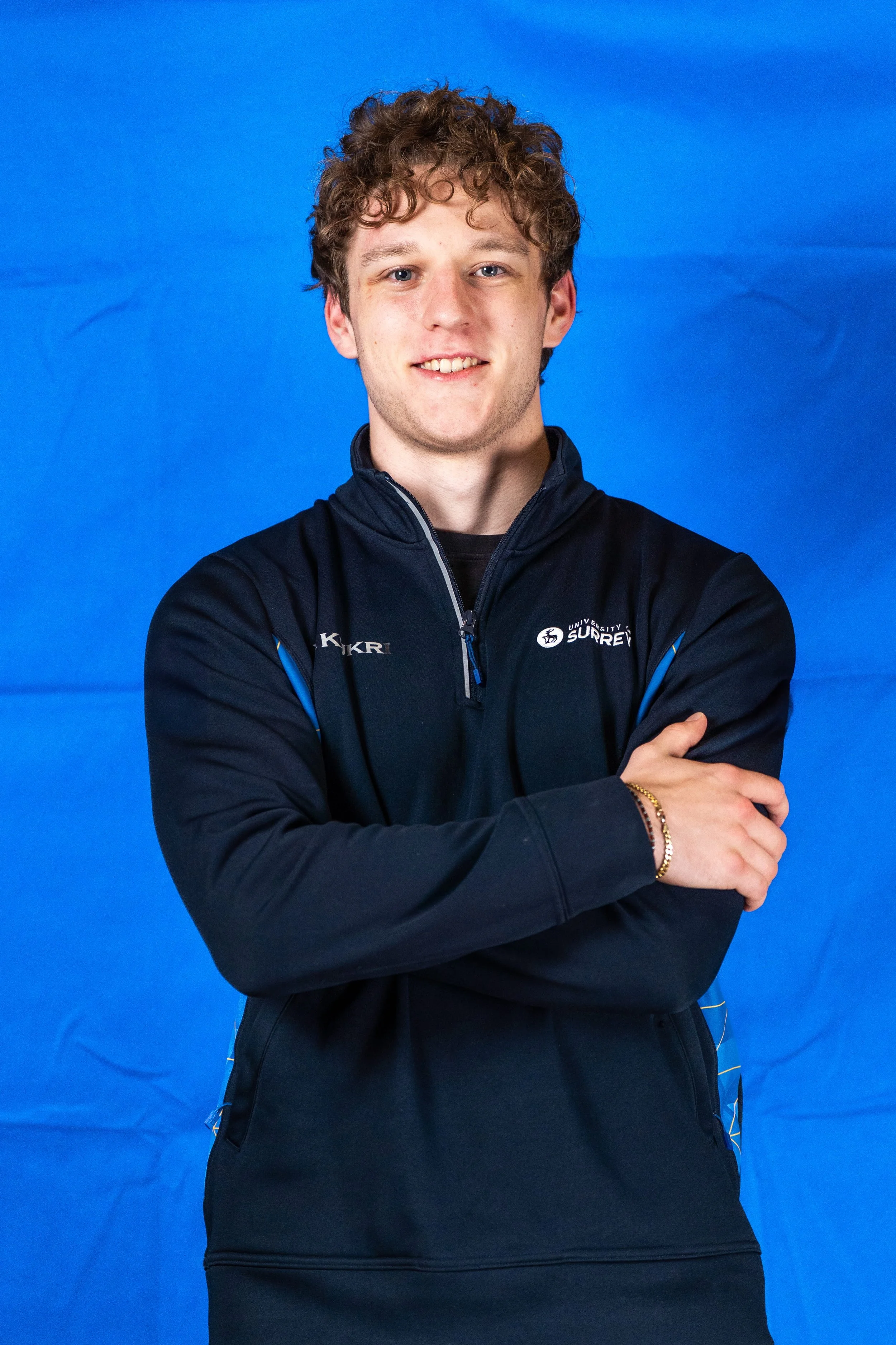 A young man with curly brown hair, blue eyes, and light skin, wearing a black University of Surrey sports jacket, standing against a blue background, smiling slightly with arms crossed.