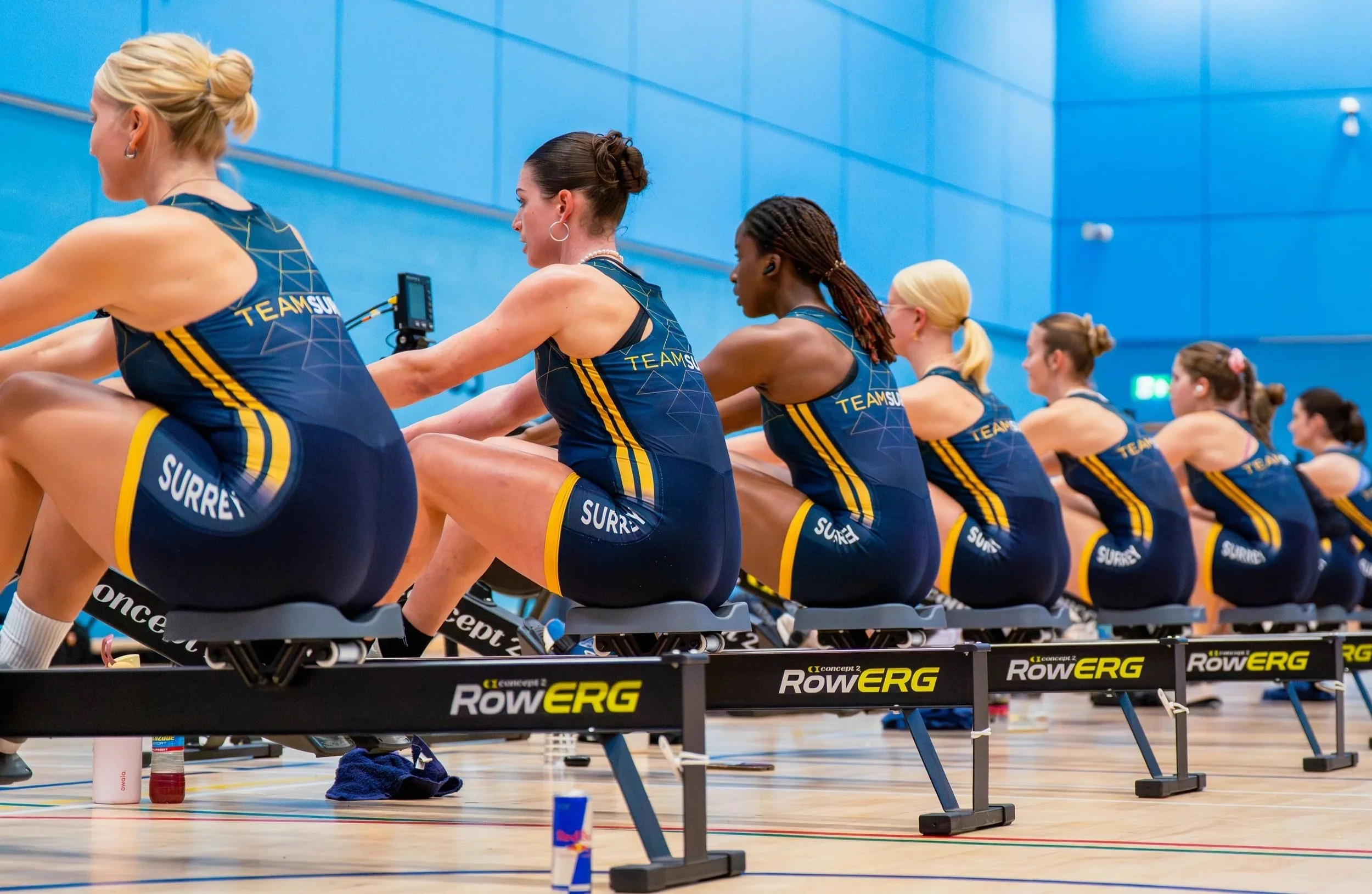Women rowers in blue team uniforms sit on indoor rowing machines in a gym, engaged in a rowing workout.