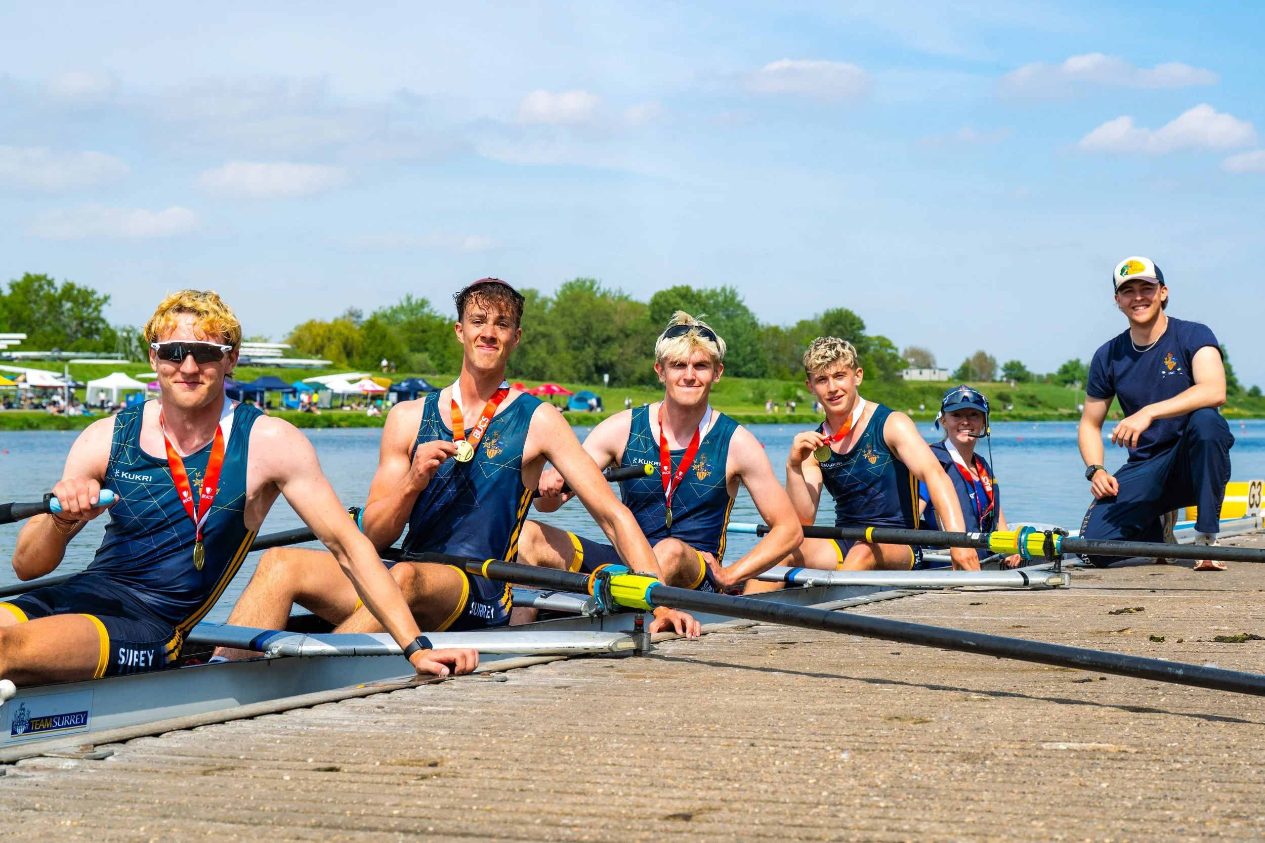 A group of young male rowers in matching sports uniforms sitting in their boat by the water, wearing medals and smiling, with a background of trees, tents, and a clear blue sky.