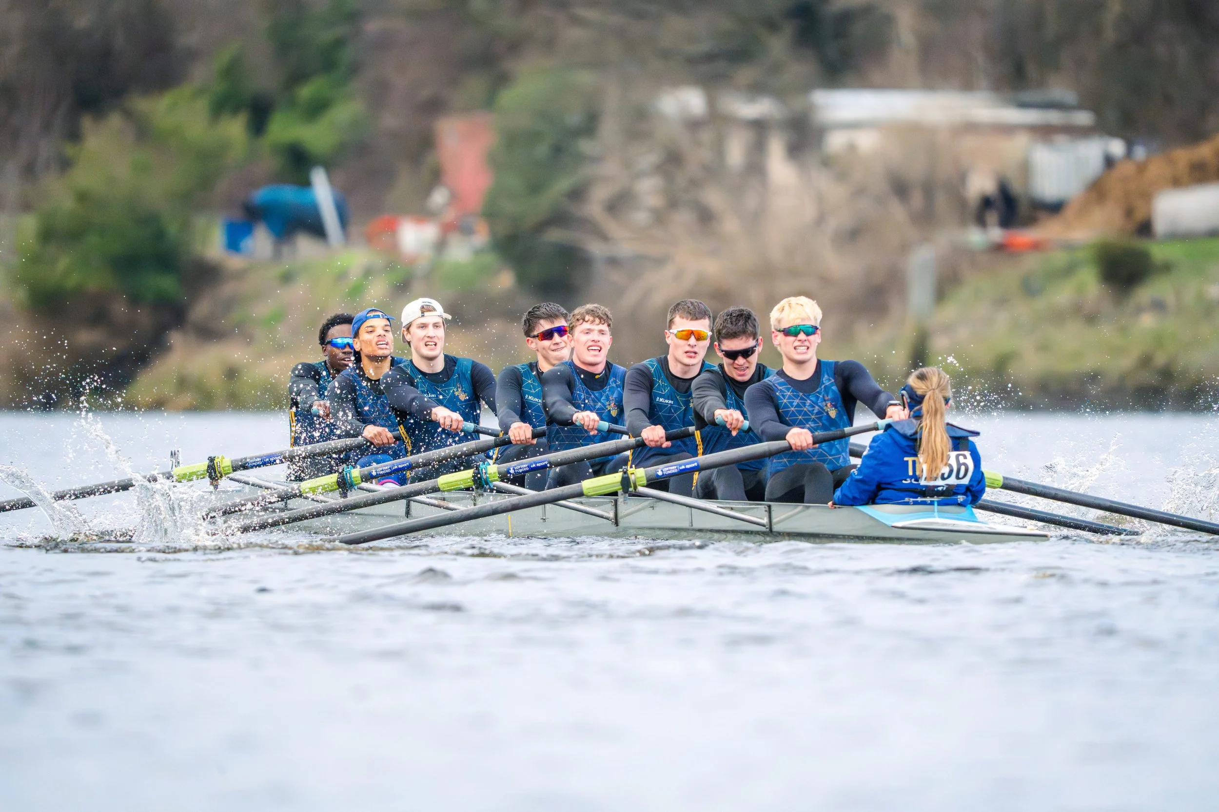 A team of eight young rowers in a racing boat on a river, wearing matching blue uniforms and sunglasses, competing intensely with their oars in the water.