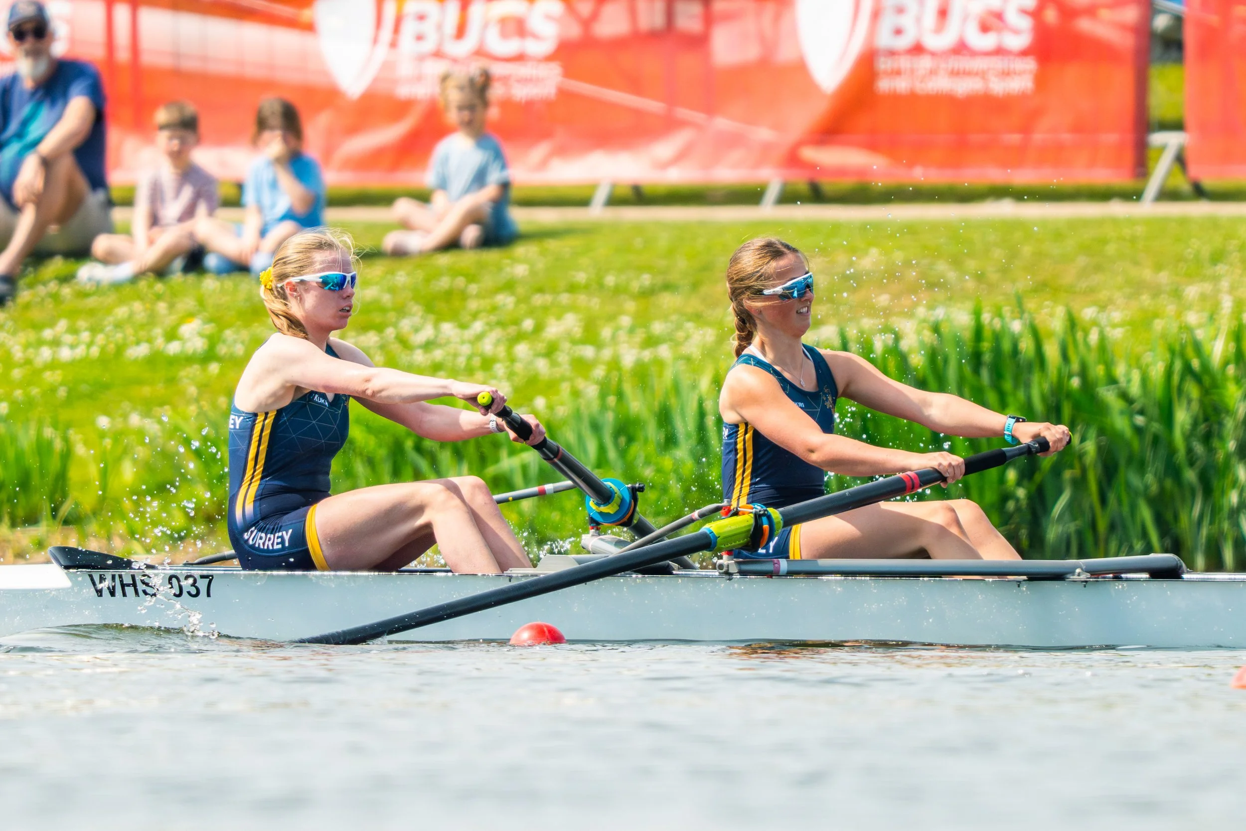 Two women wearing athletic gear and sunglasses are paddling in a double kayak on a body of water during a race, with a red buoys marking the course and a grassy area with spectators in the background.