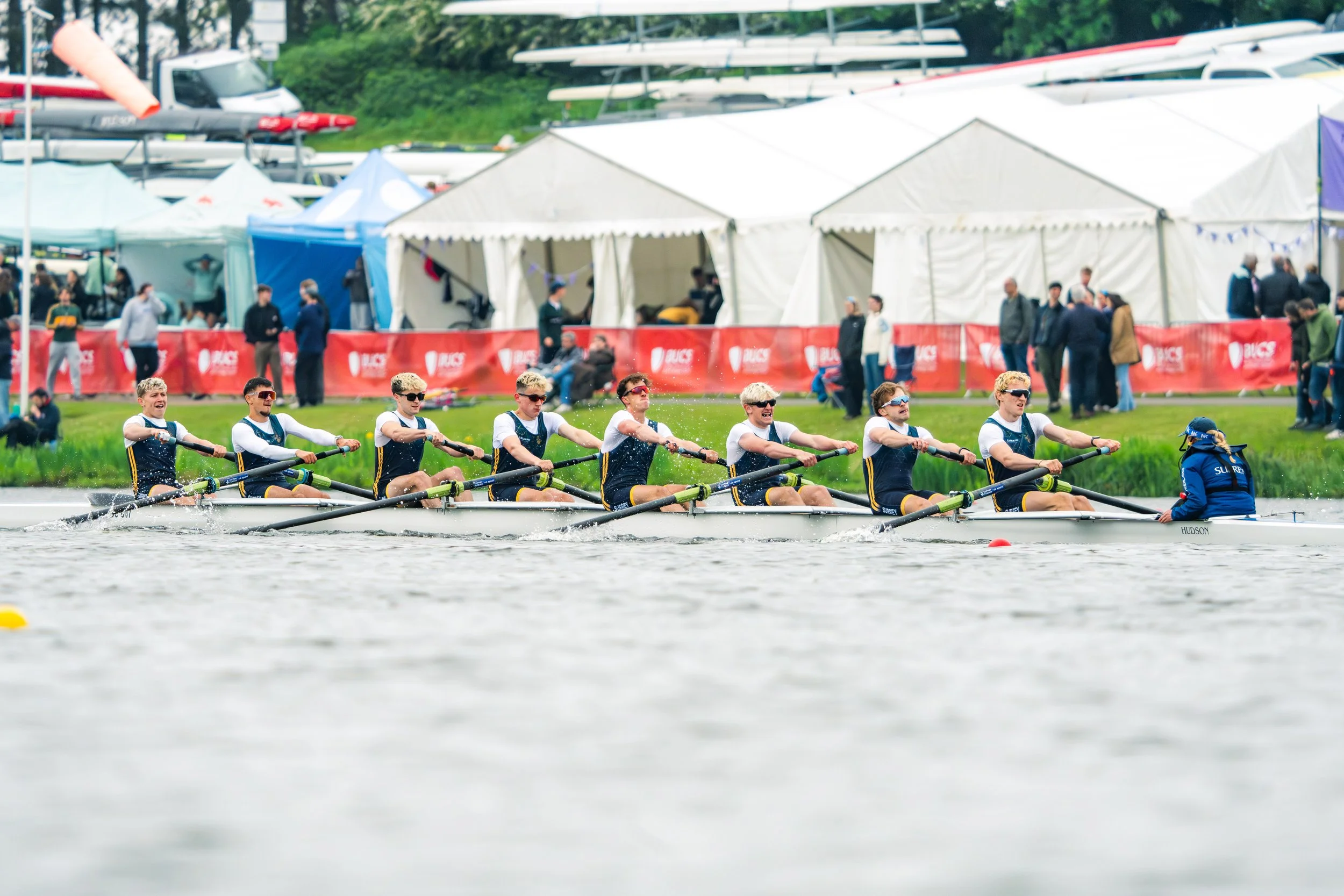A team of young rowers wearing uniforms and sunglasses competing in a rowing race on a river, with tents and spectators in the background.