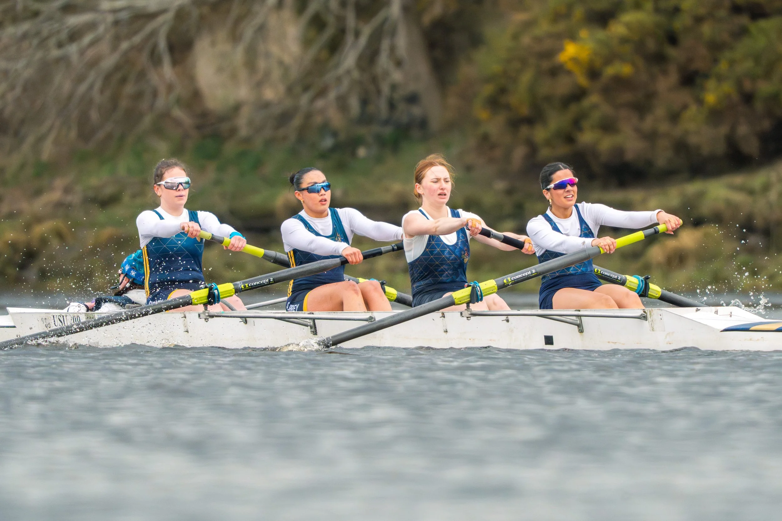 A team of four women rowing a boat on a body of water during daytime, with trees in the background.