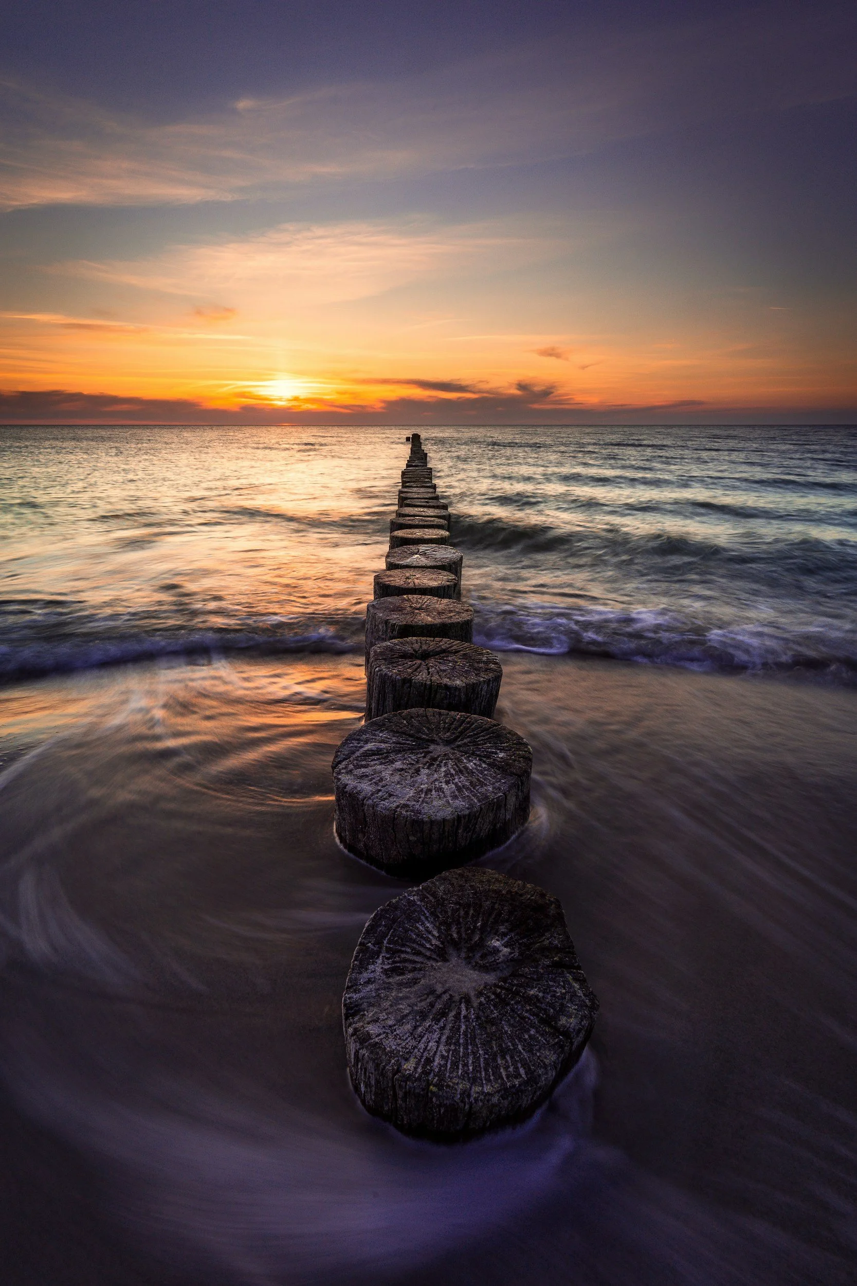 A wooden pier extending into the ocean during sunset with a colorful sky and gentle waves.