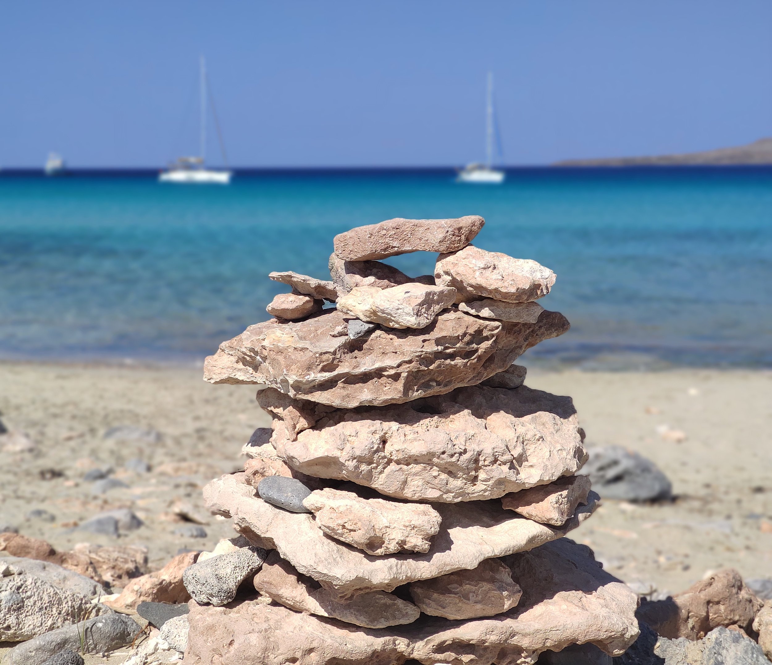 A stack of flat rocks balanced on each other on a beach, with the ocean and sailboats in the background under a clear blue sky.