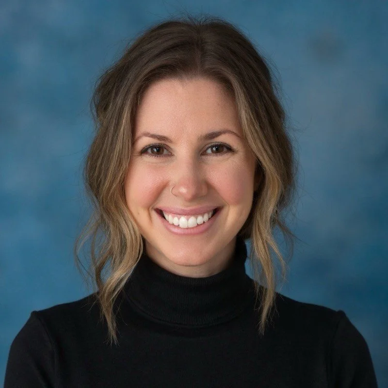 A woman with shoulder-length wavy brown hair, wearing a black turtleneck, smiling in front of a blue background.