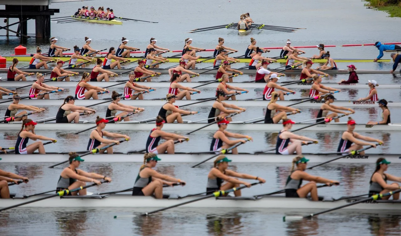 Starting line of a 6 boat heat at the NCAA women's rowing championships.  The rowing race is on a lake, with teams in colorful uniforms and hats, each in a women's eight oared shell.