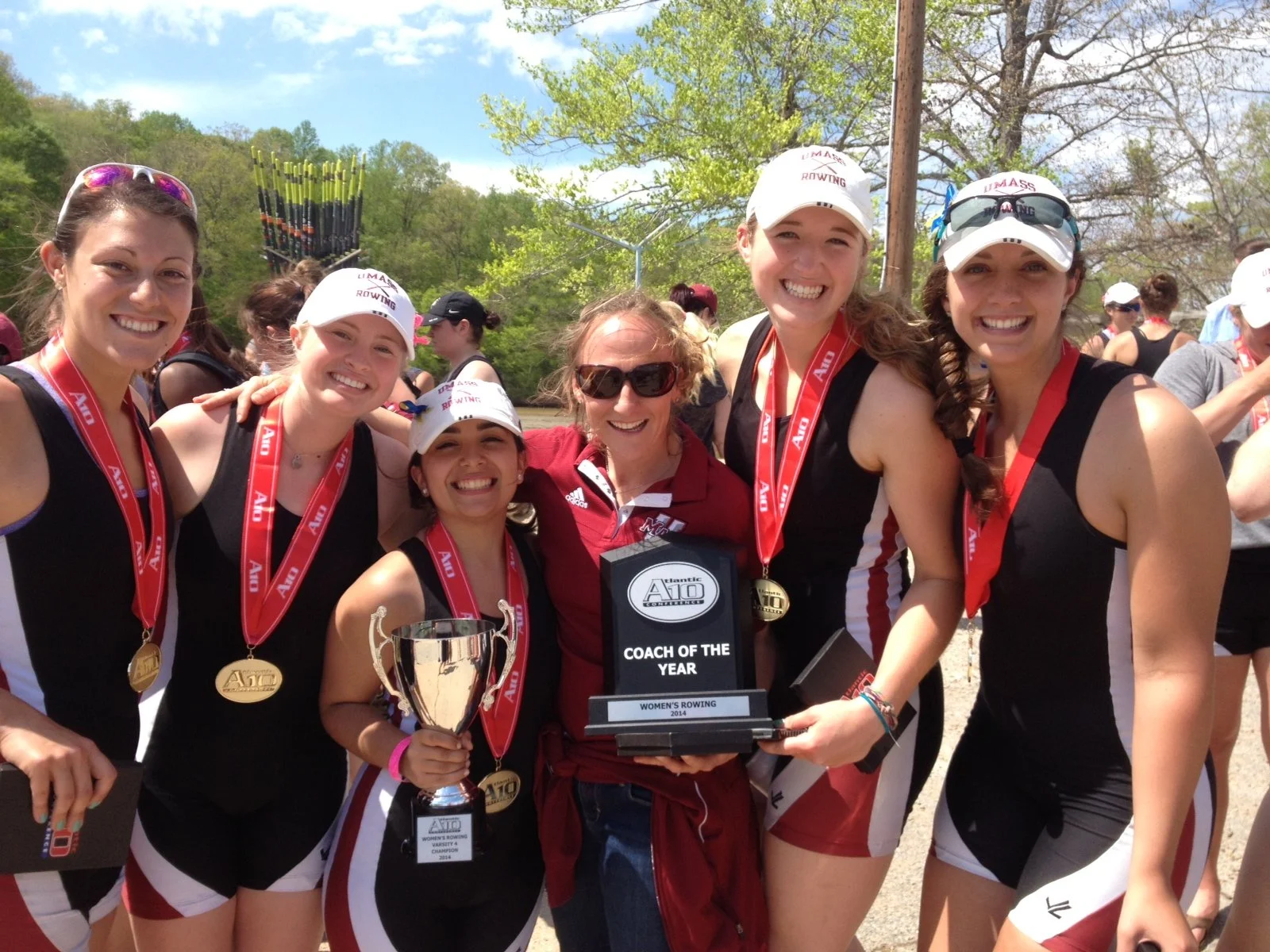 Group of female UMass rowers with medals and their coach, Laura Simon, holding a trophy and a plaque that reads 'Coach of the Year' from Atlantic A10 Conference, celebrating after the A-10 Conference Championships.