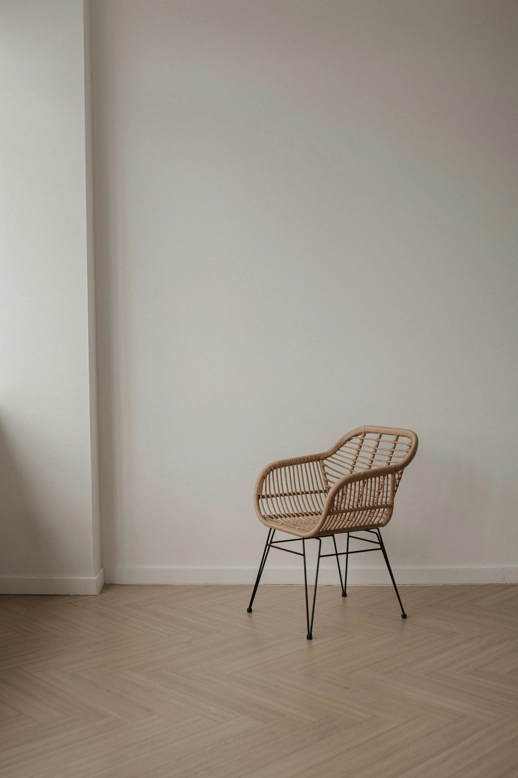 A rattan chair with black metal legs placed against a plain, light-colored wall and light wood flooring.
