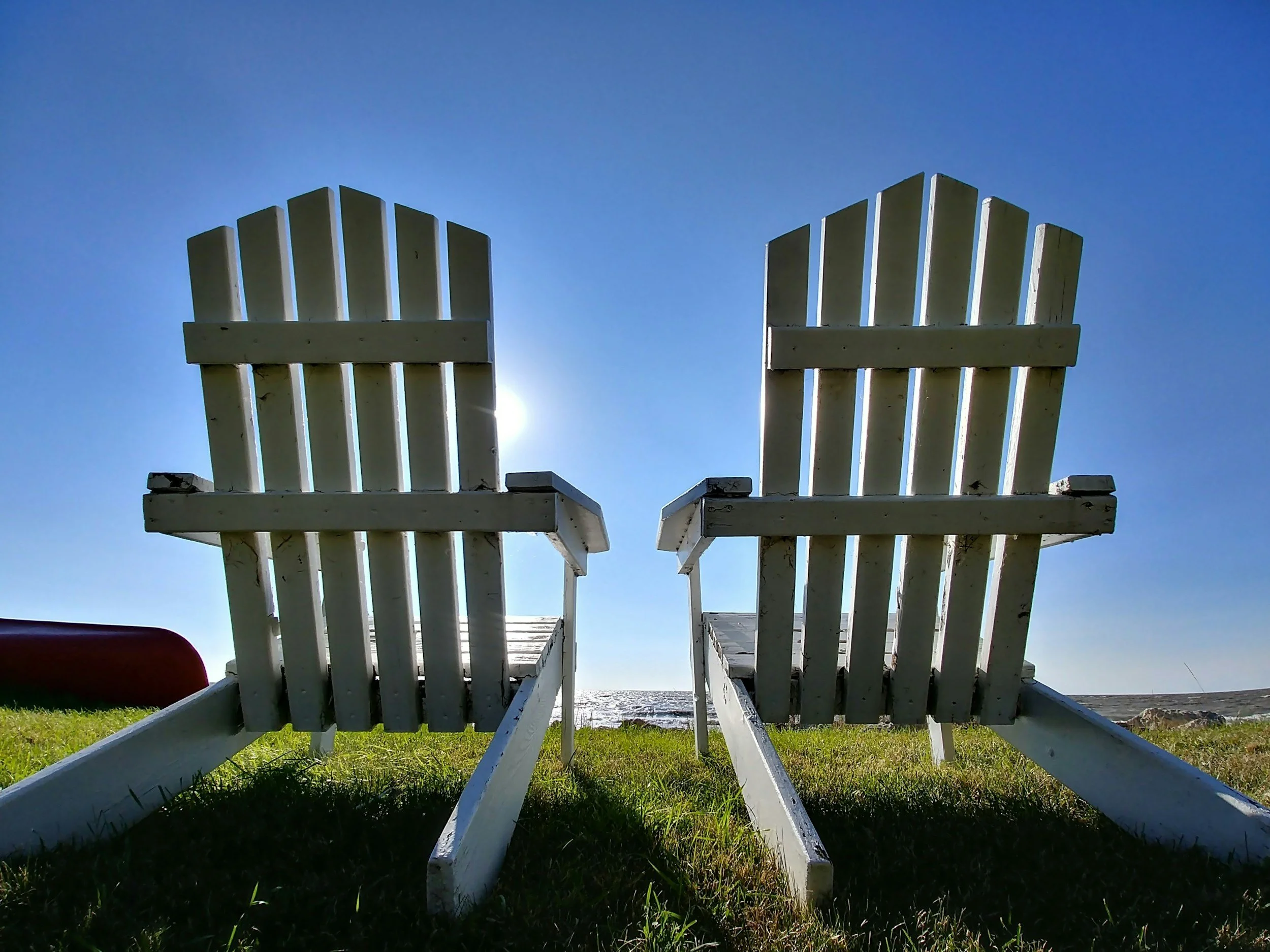 Two white Adirondack chairs facing the ocean on a grassy beach, with a blue sky and the sun in the background.