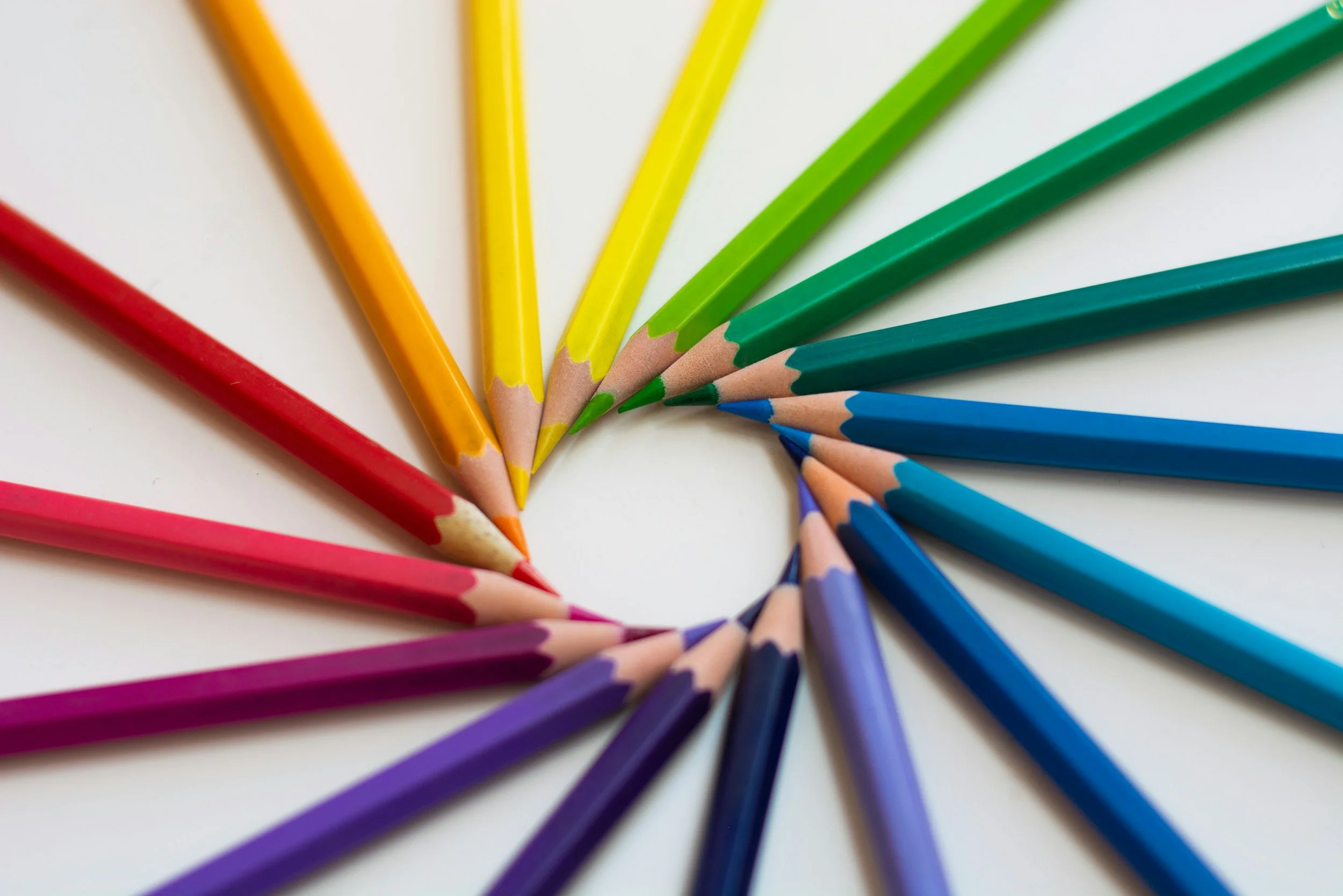 A close-up of colored pencils arranged in a fan shape on a white surface, displaying various colors like red, orange, yellow, green, blue, and purple.