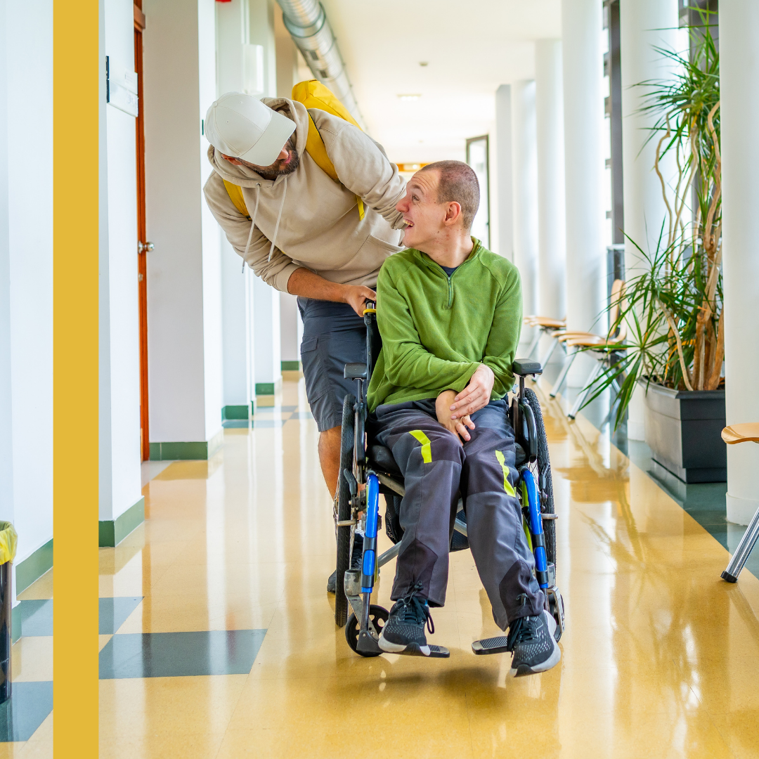Two people in a corridor, one seated in a wheelchair, both smiling and leaning towards each other in conversation.