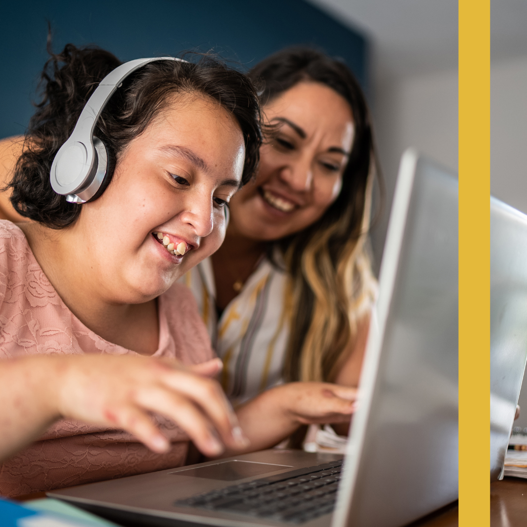 Two people smiling together at a laptop, one wearing headphones.