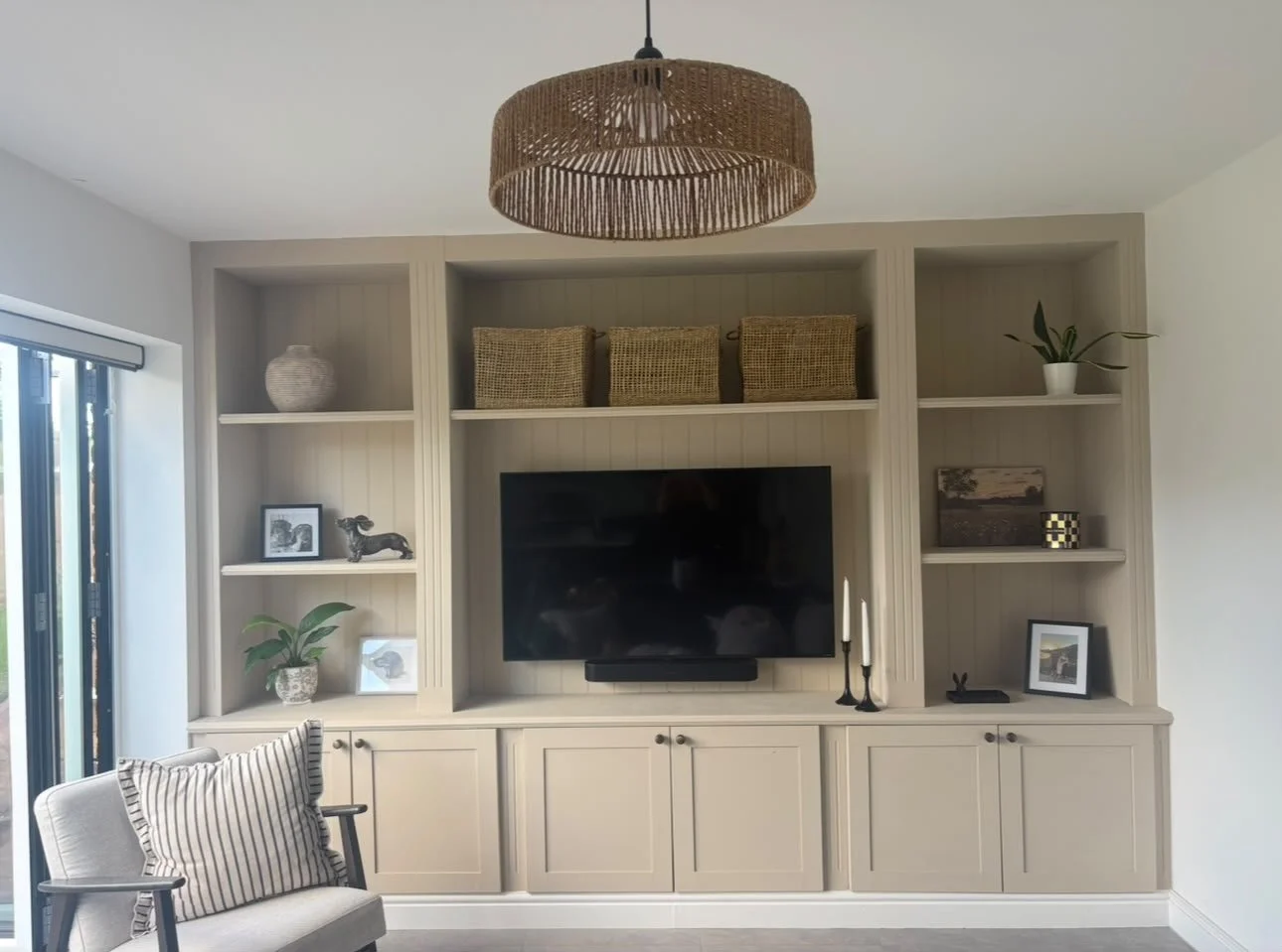 Living room built-in entertainment center with shelves, a flat-screen TV, decorative baskets, framed photos, potted plants, and candles, next to a light-colored armchair with striped pillow.