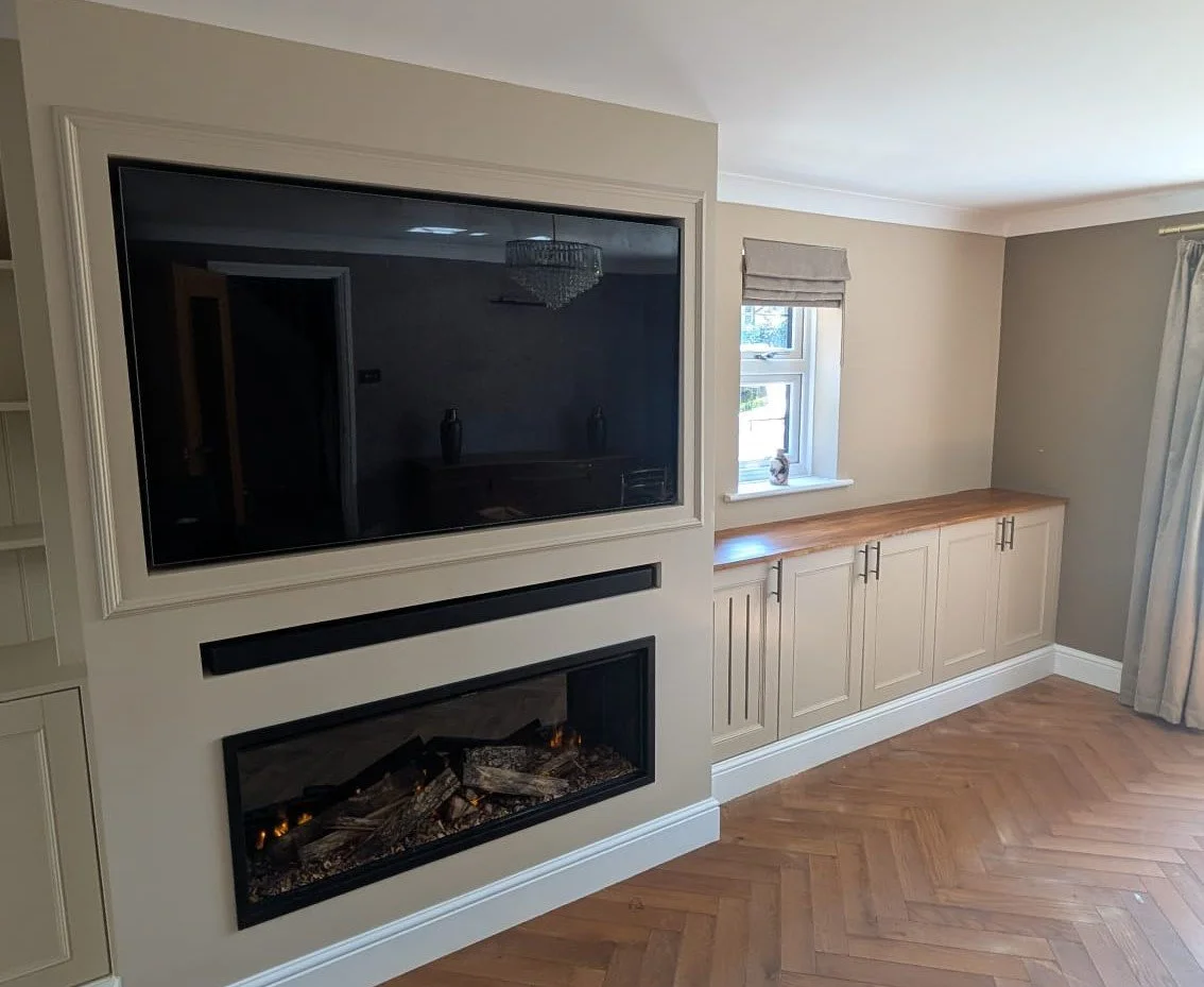 Living room with a built-in entertainment center and an electric fireplace, with a window and cabinet beneath, beige walls, hardwood floor, and curtains.