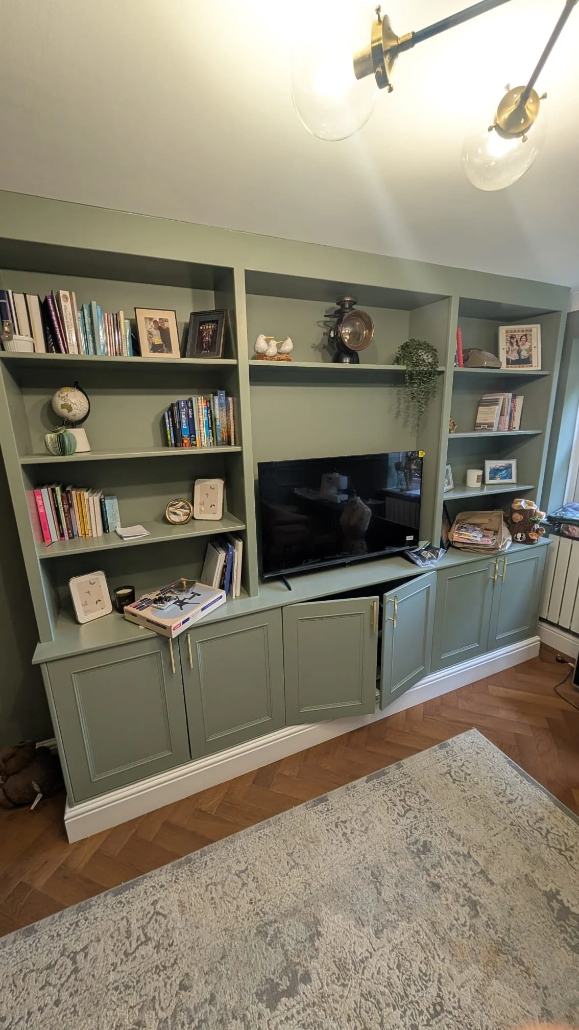 A green wooden entertainment center with books, family photos, and decorative items, including figurines and framed pictures. There is a flat-screen TV and various items on the shelves and cabinets. The room has hardwood floors and a light-colored rug.