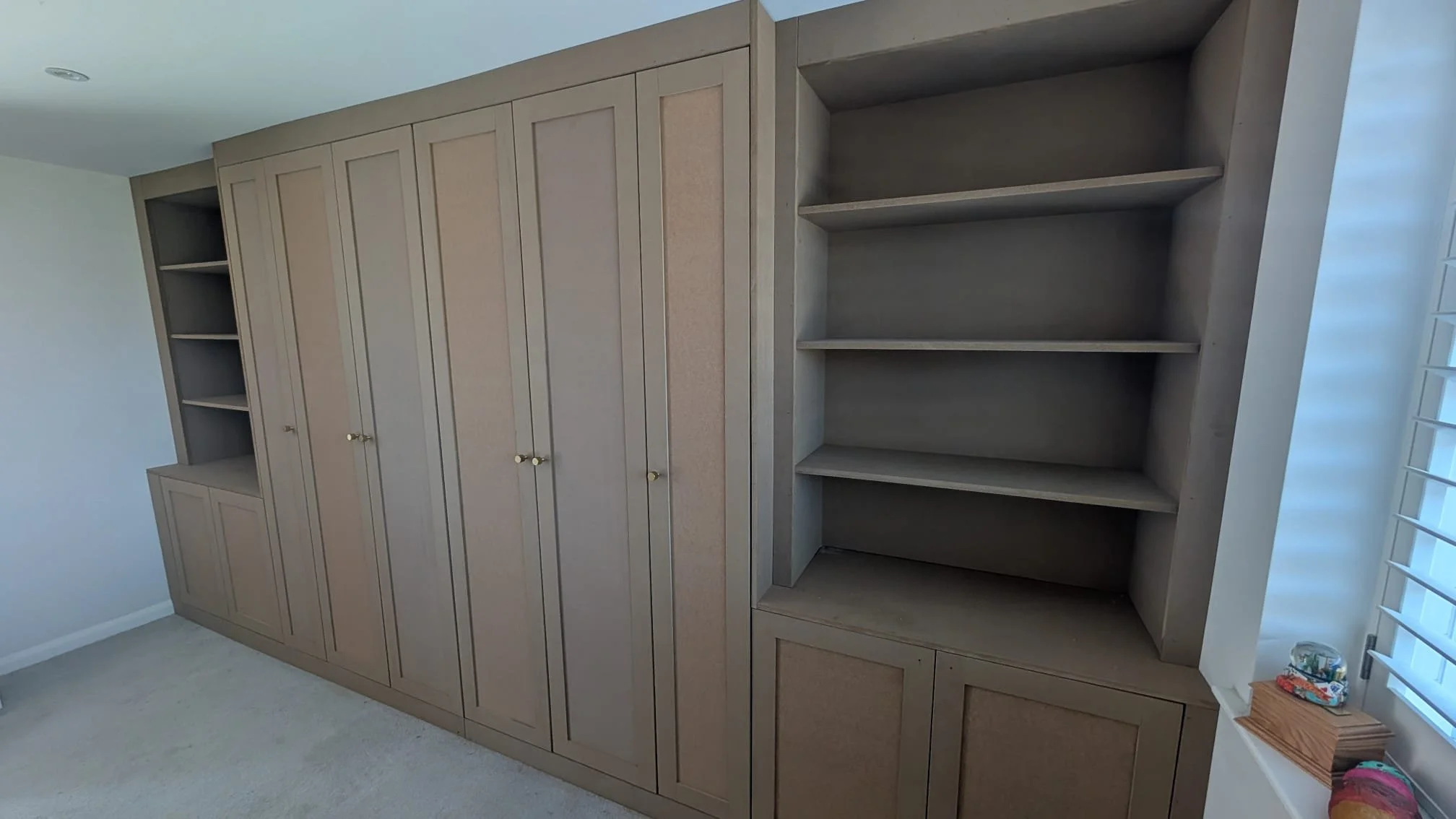 A large beige built-in bookshelf with multiple closed cabinets at the bottom and open shelves at the top, situated near a window with white blinds.