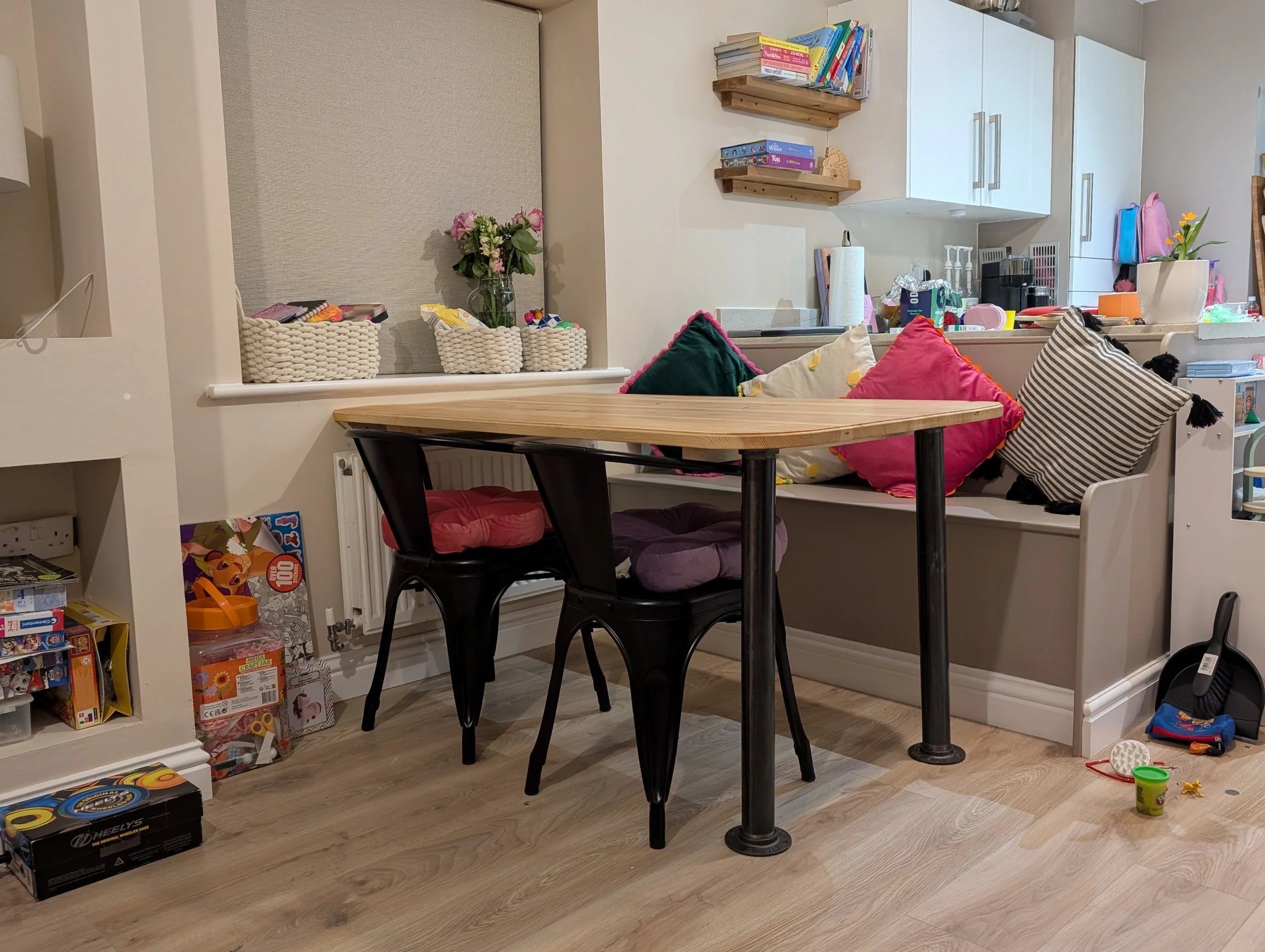 Living room with a wooden dining table surrounded by three dark chairs, colorful pillows on a bench, toys and books scattered on the floor, a kitchen area with cabinets, shelves with books, plants, and various kitchen items, and a window with a beige roller blind.