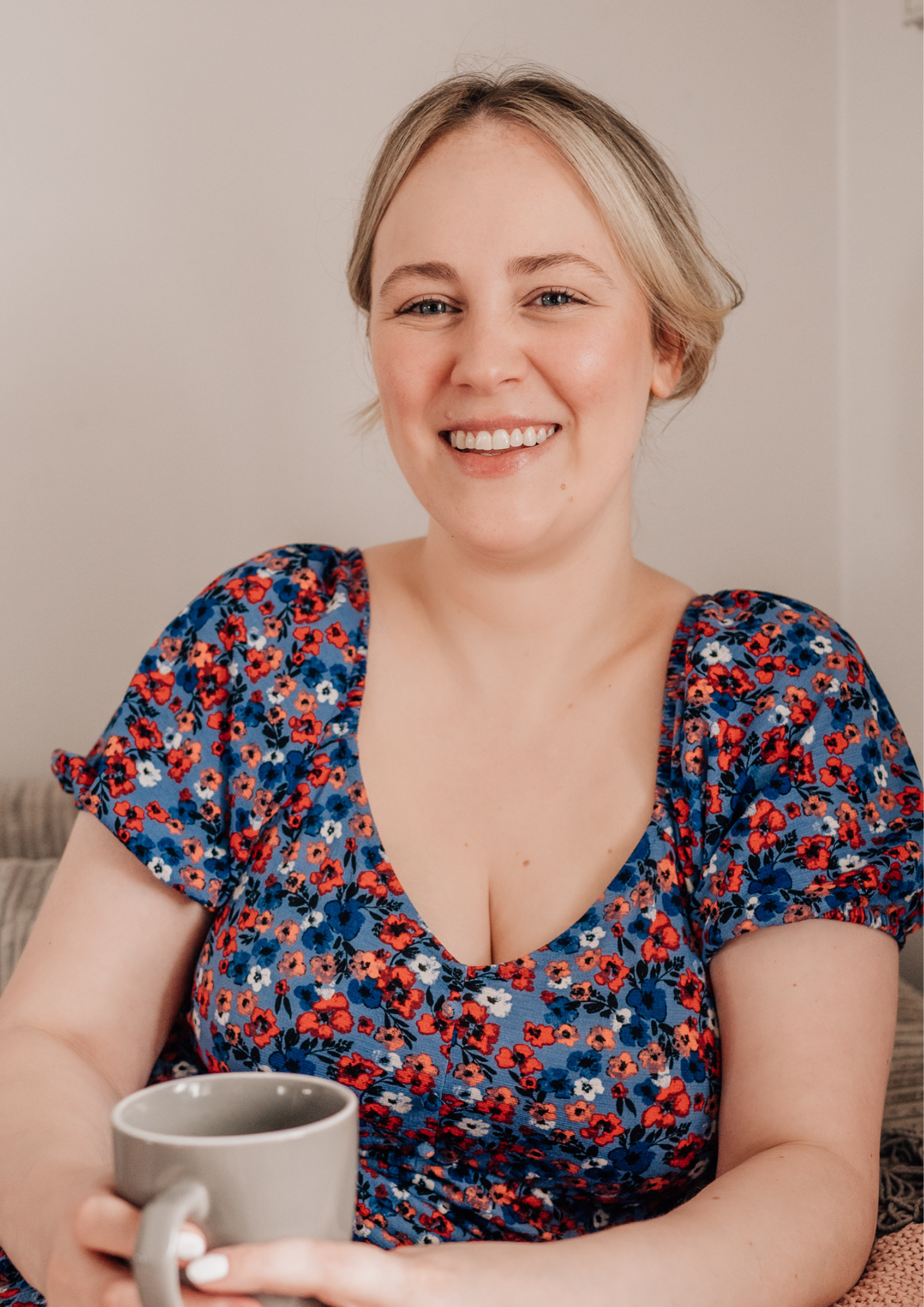A woman with short blonde hair wearing a colorful floral dress holding a gray mug, smiling at camera.