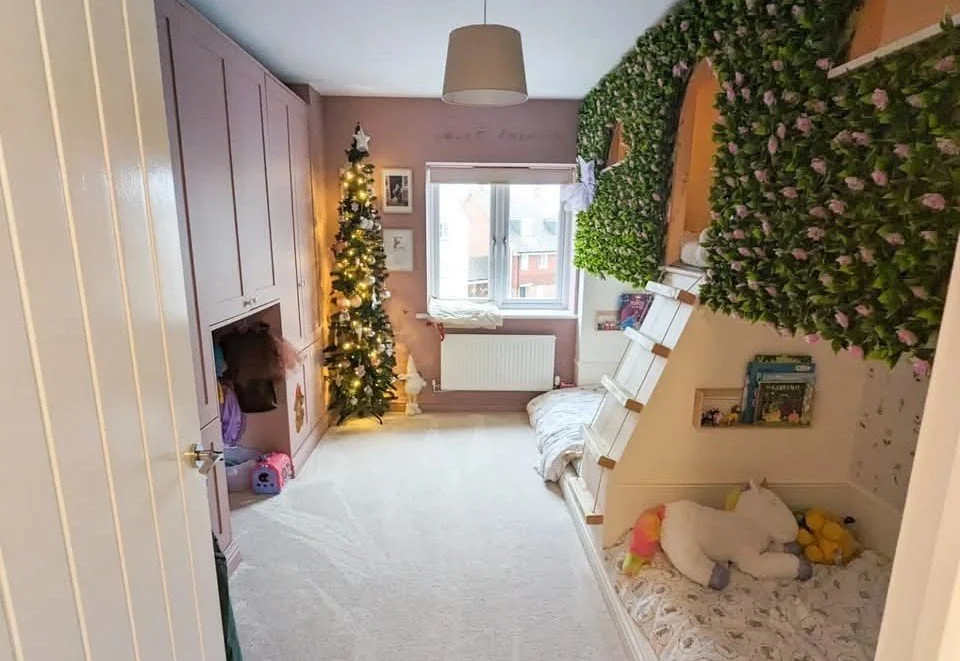 A child's bedroom decorated for Christmas with a small lit Christmas tree, a window with books and toys nearby, purple wardrobe, pink walls, and a cozy corner with stuffed animals and a small bed.