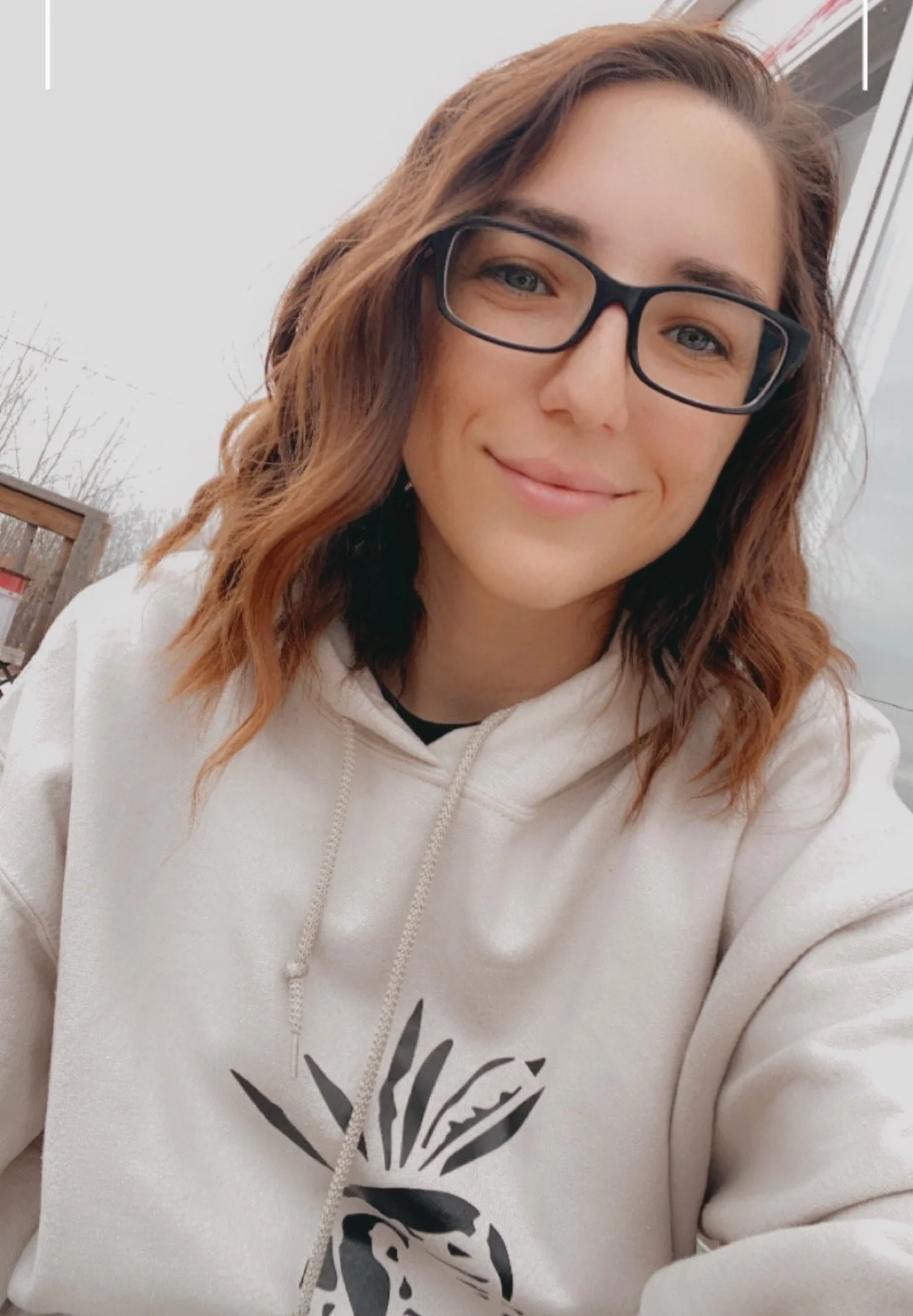 A woman with wavy, shoulder-length auburn hair, wearing glasses and a beige hoodie with a zebra print on the front, smiling outdoors on a cloudy day.