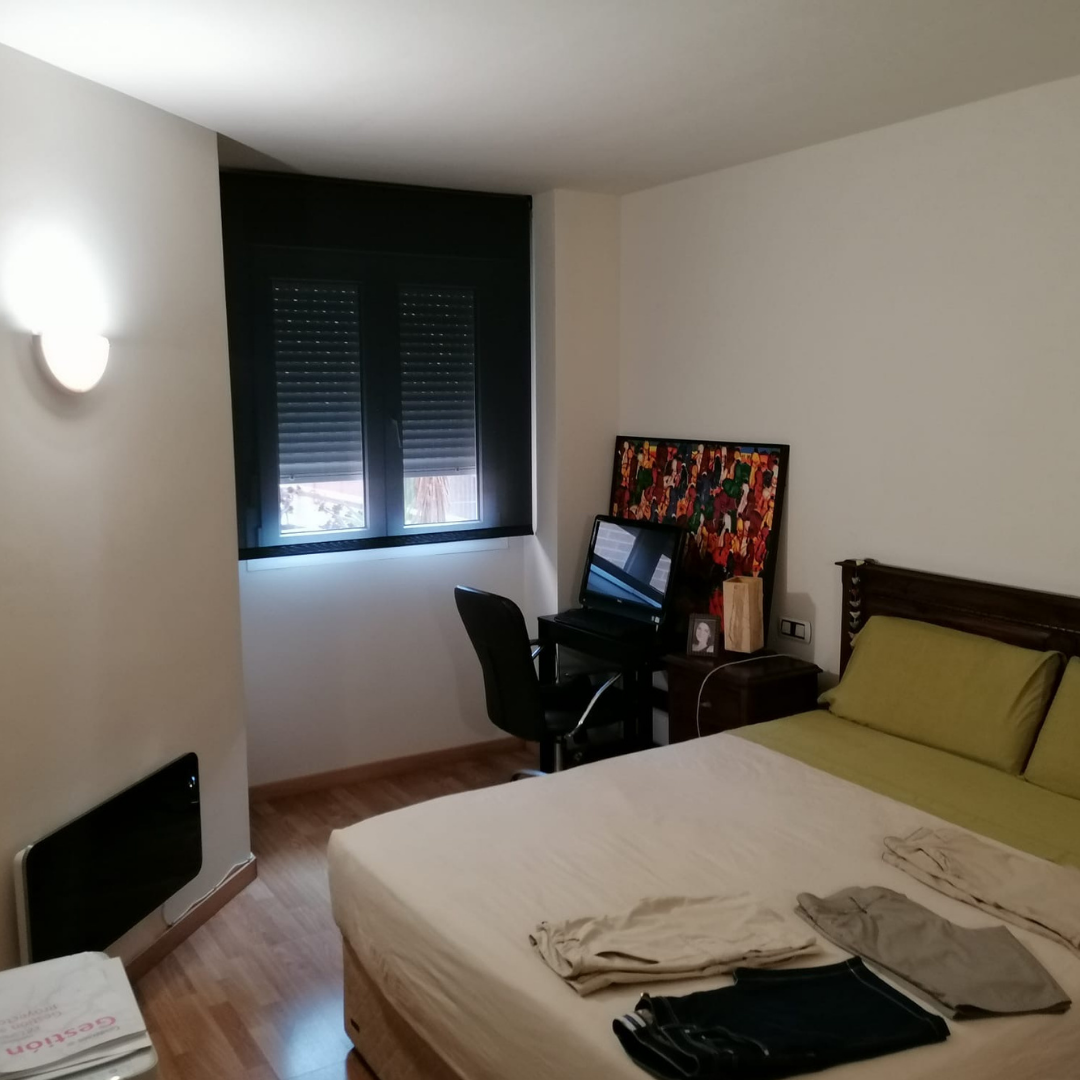 Habitación con cama, escritorio con computadora y silla, ventana con cortinas negras, paredes blancas, luz de pared, piso de madera, ropa en la cama, una televisión en el suelo.