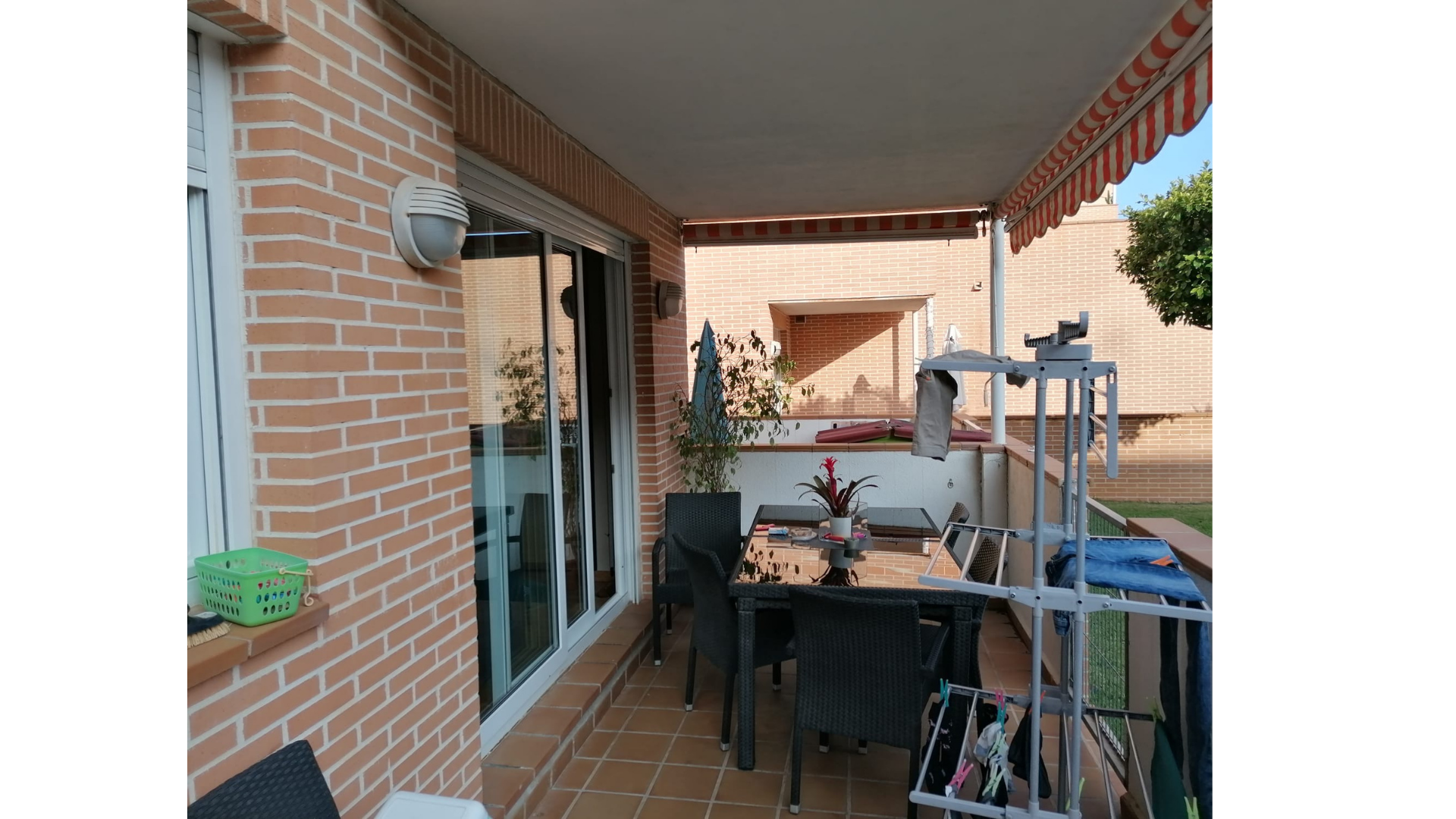 Balcón con mesa y sillas, plantas, ropa colgada, toldos y paredes de ladrillo.