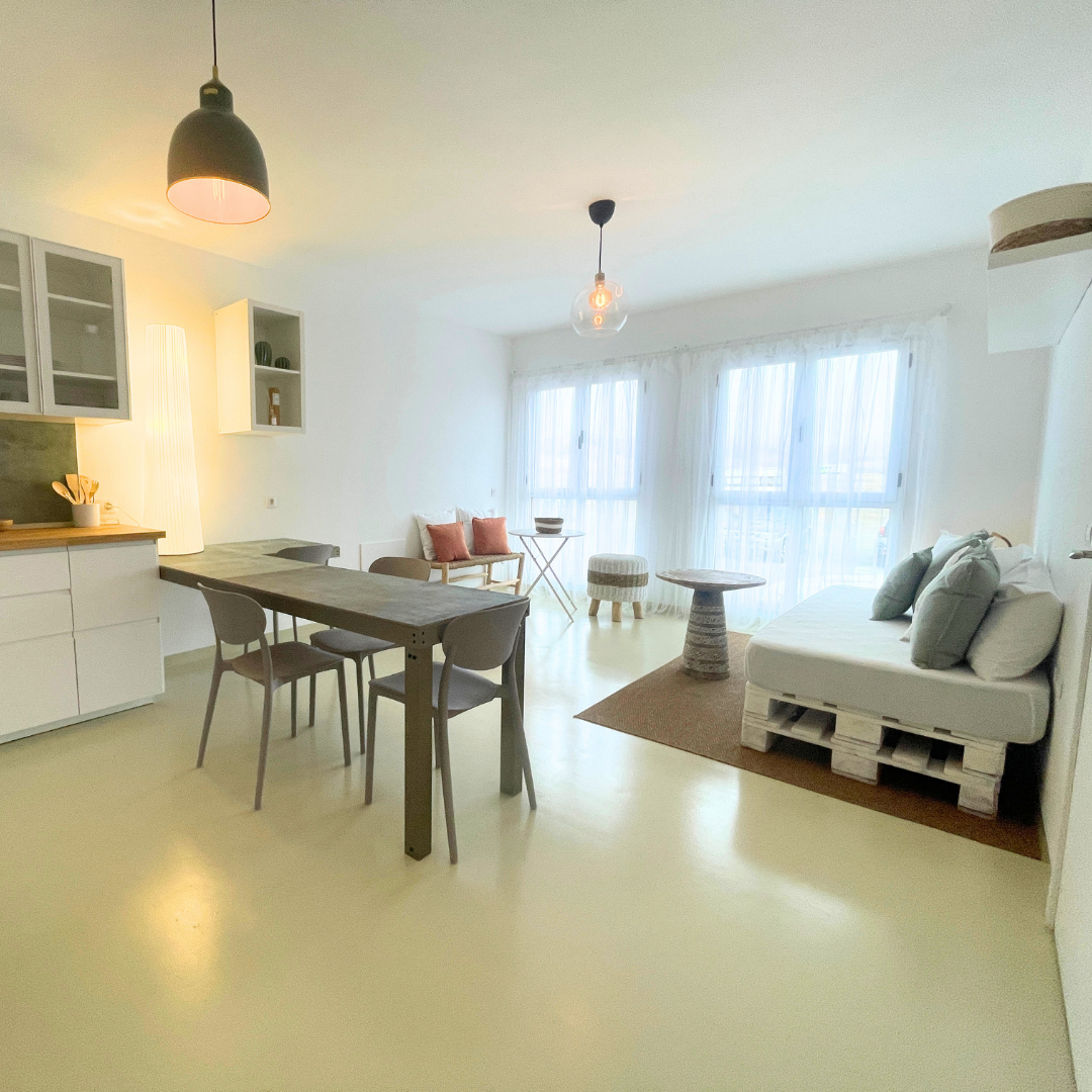 Sala de estar con sofá de palets, mesa de madera, sillones pequeños, cortinas blancas y lámparas de techo.