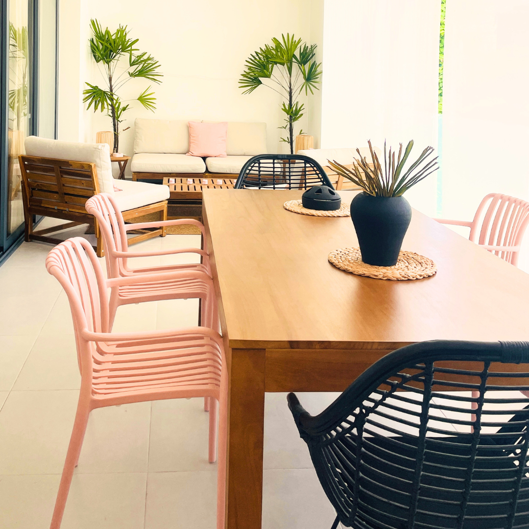 Sala de estar con sillones blancos, plantas grandes, mesa de comedor de madera con sillas rosas y negras, y decoración de plantas en jarrones.