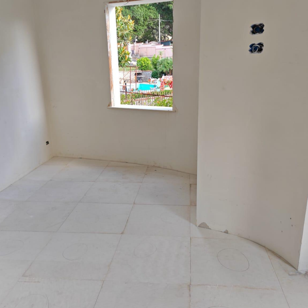 Habitación en construcción con piso de baldosas blancas, una ventana sin marcos y paredes blancas, una de las paredes tiene cables eléctricos expuestos.