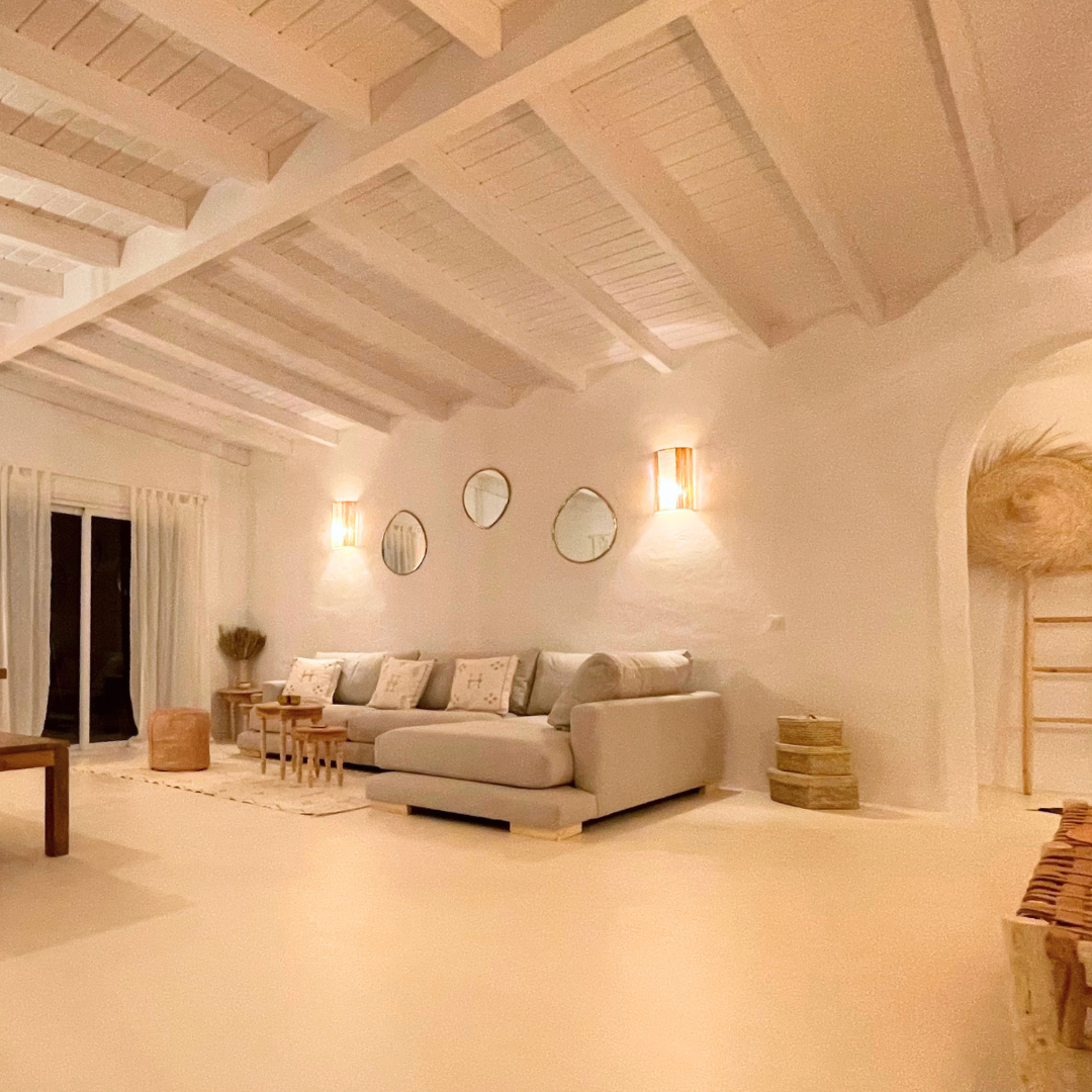 Sala de estilo mediterráneo con muro y techo blancos, sofá gris claro con cojines, espejos en la pared, luminarias, una mesa baja y decoraciones en tonos neutros.