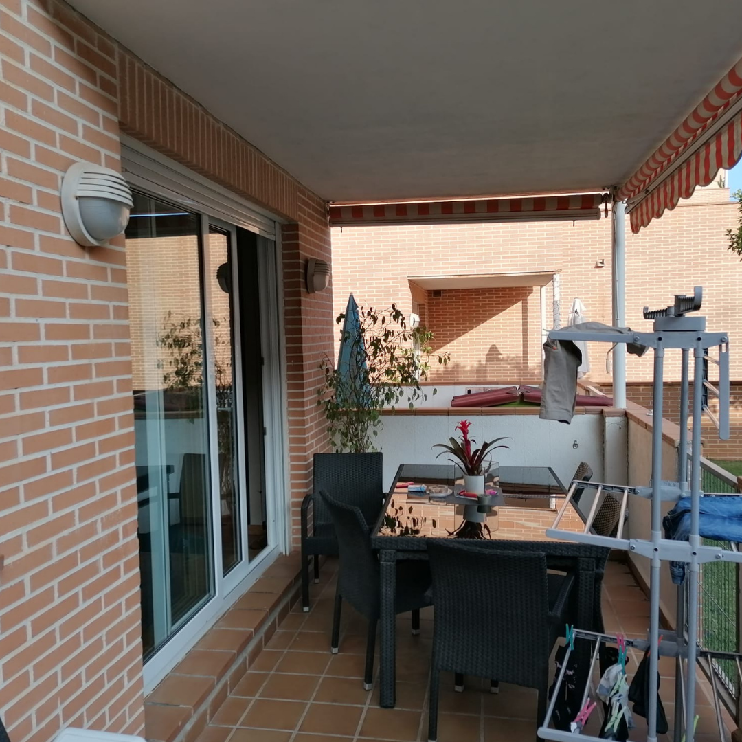 Terraza con mesa de cristal, sillas negras, plantas y estructura para secar ropa, rodeada de paredes de ladrillo y con toldo rojo y blanco.