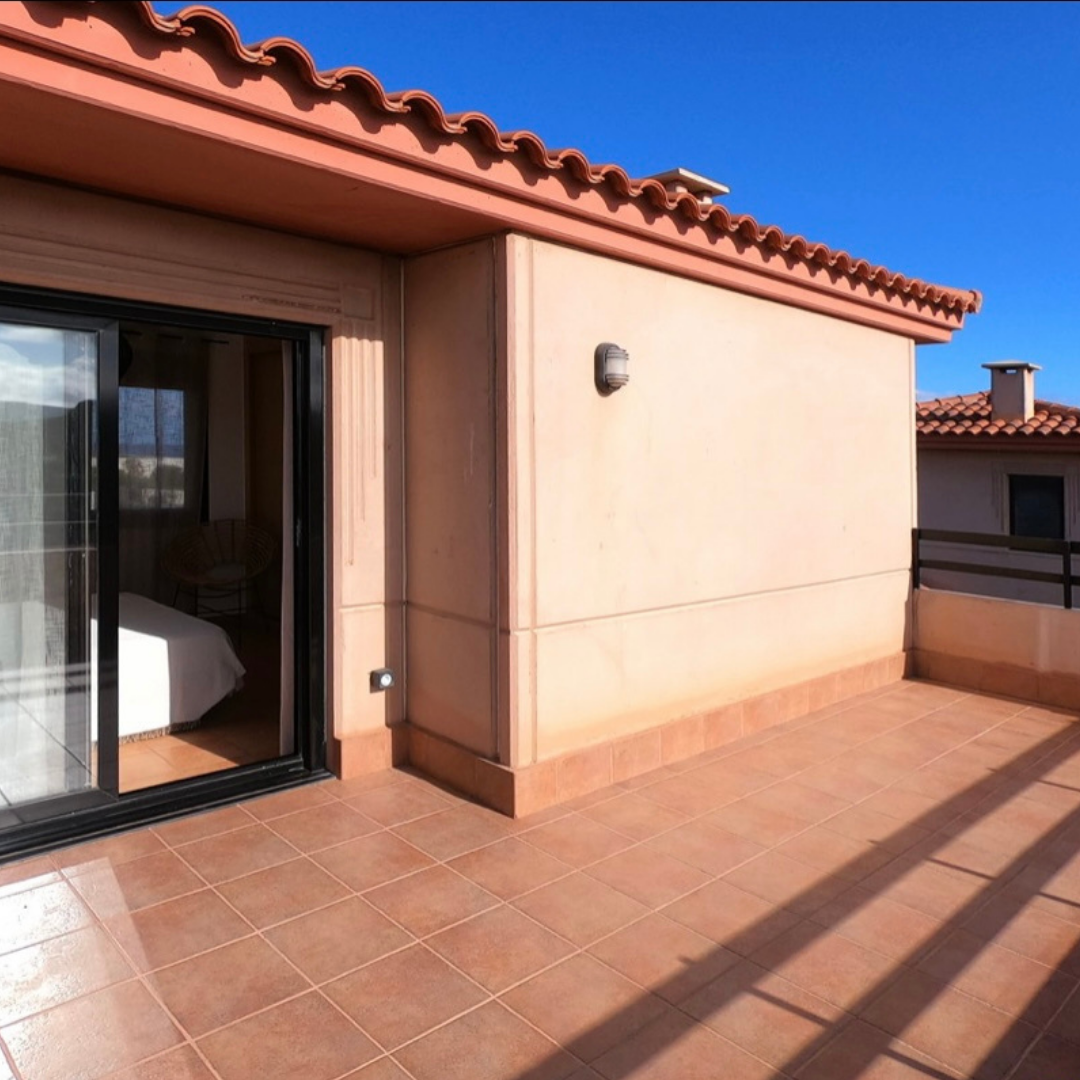 Terraza de azulejos en un apartamento con pared beige, ventanales corredizos y cielo azul.