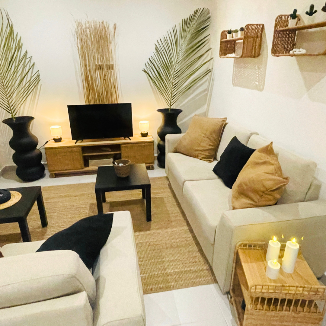 Sala de estar con muebles blancos, cojines en tonos beige y negro, mesa de centro negra, televisión en mueble de madera, lámparas, plantas grandes en macetas negras, estantes de madera en la pared con plantas pequeñas y decoraciones, alfombra de fibra natural, velas en una mesa de mimbre.