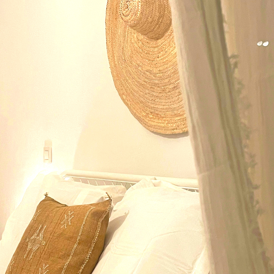 Cama con almohadas blancas y cojín de tela marrón, pared blanca, sombrero de paja colgado en la pared, interruptor de luz, superficie de madera en primer plano