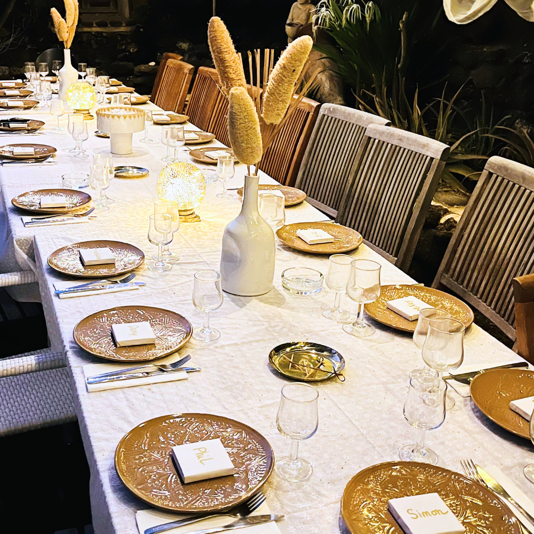Mesa decorada para una comida o cena, con un mantel blanco, platos dorados, copas y cubiertos, y un centro con un jarrón blanco y vegetación, en un ambiente elegante y bien iluminado.