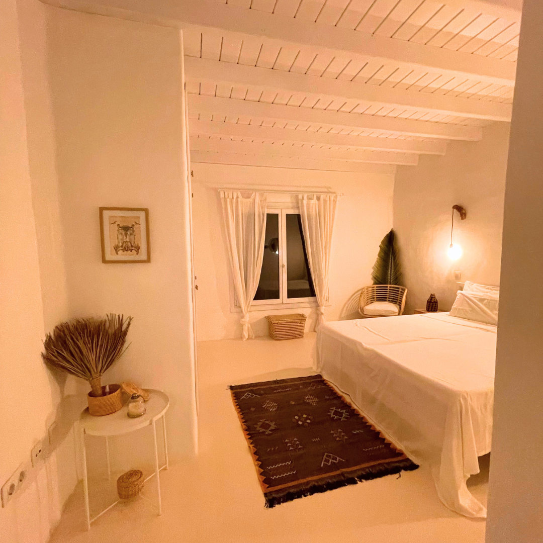Habitación con paredes claras, techo de madera blanca, cama con ropa blanca, alfombra negra con diseños, ventana con cortinas blancas, silla de mimbre, lámpara colgante y decoración con plantas y cuadros.