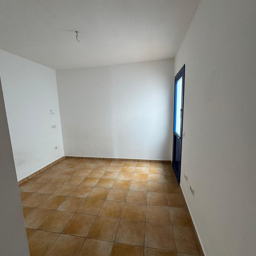 Una habitación vacía con paredes blancas, suelo de baldosas marrones y una puerta azul hacia afuera.