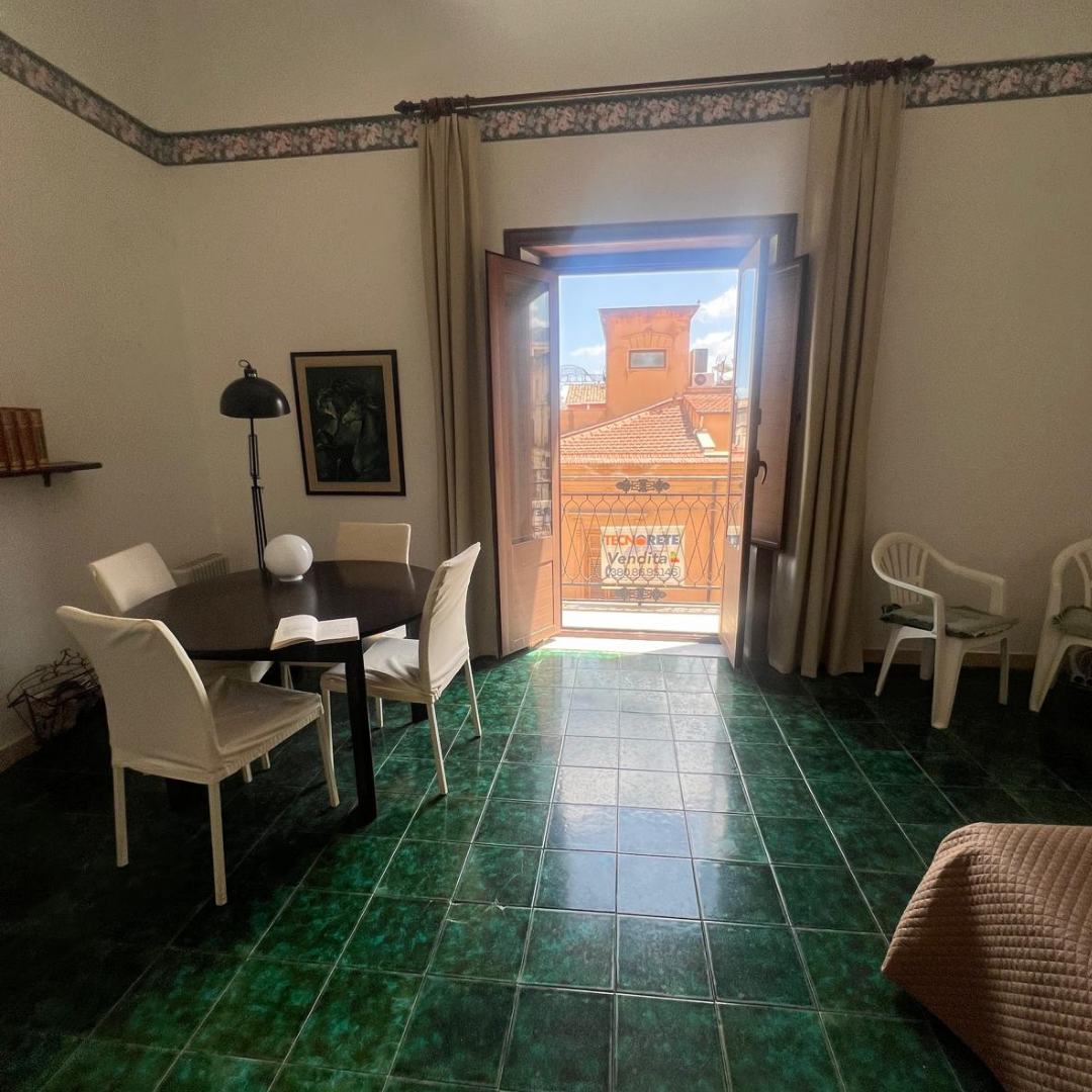Sala con sillones blancos, mesa redonda con sillas, puertas abiertas a un balcón con vista a tejados y un edificio, piso de baldosas verdes.