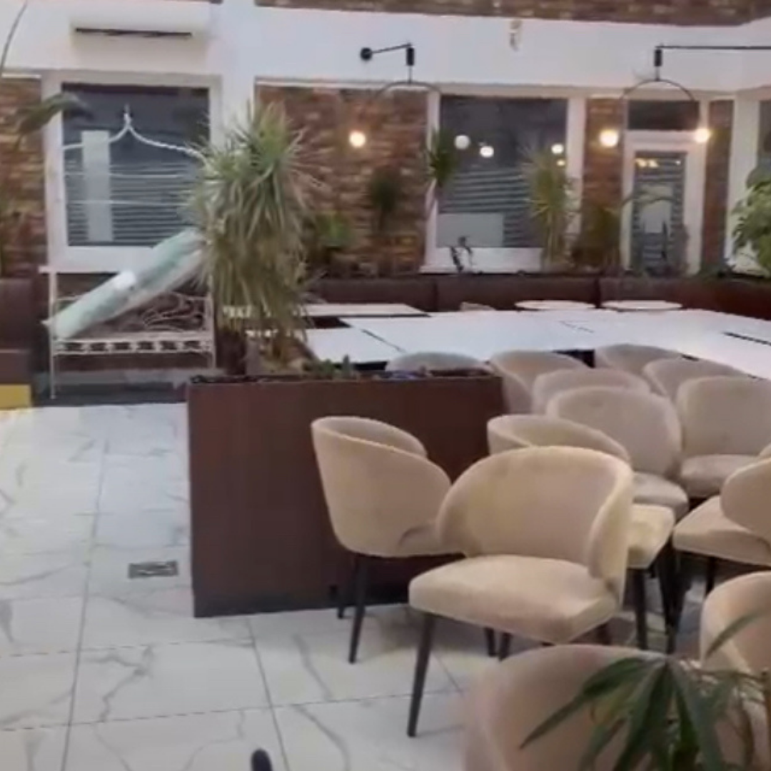 Área de comedor al aire libre con sillas beige, mesas blancas y plantas decorativas, con paredes de ladrillo y ventanas con persianas.